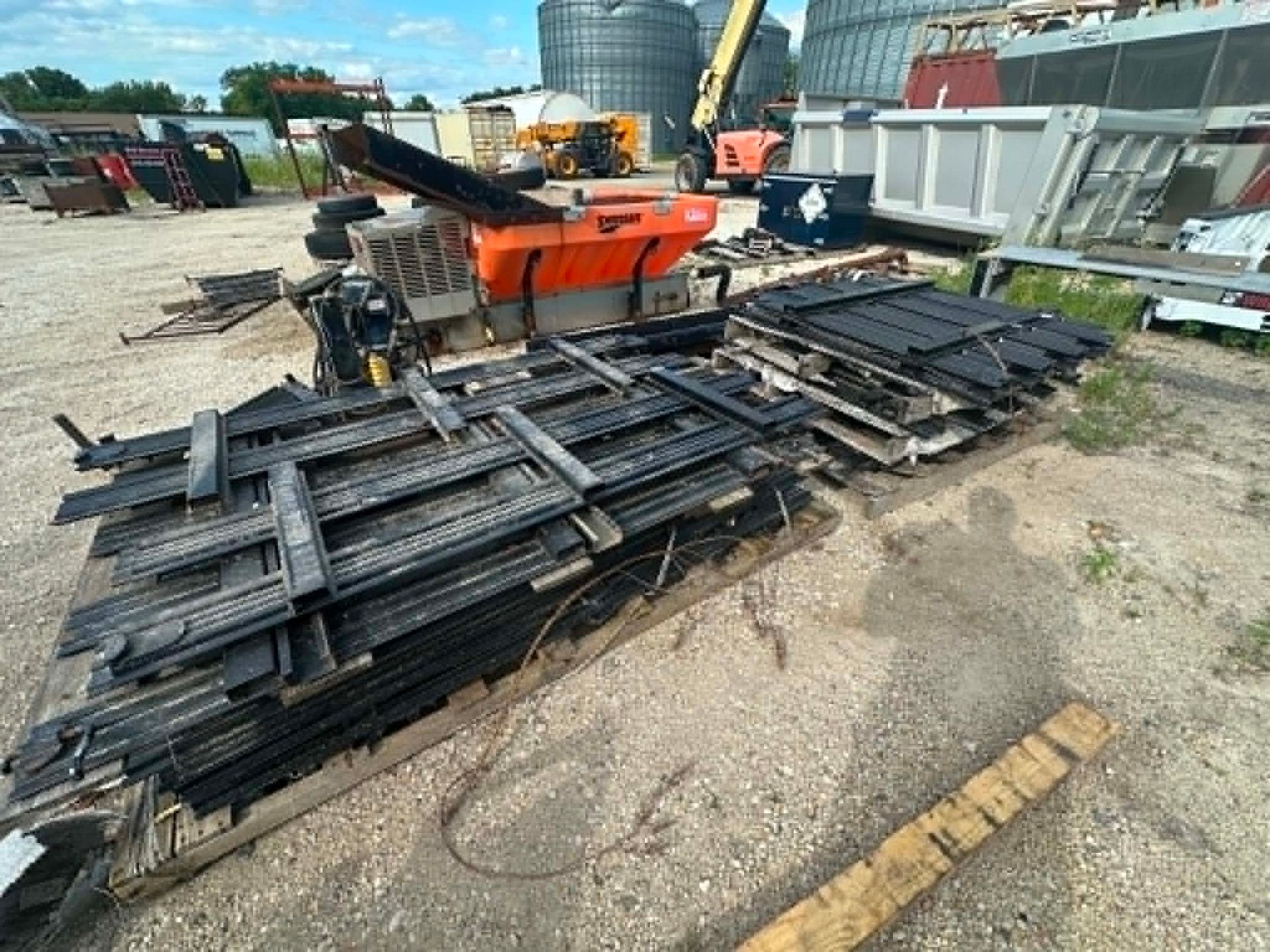 Stacked black metal beams beside an orange industrial machine in an outdoor scrapyard.