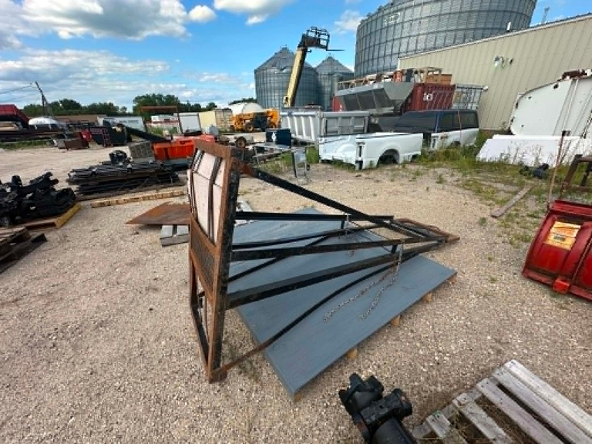 Damaged metal billboard frame leaning on gravel in an industrial yard, with vehicles and equipment nearby