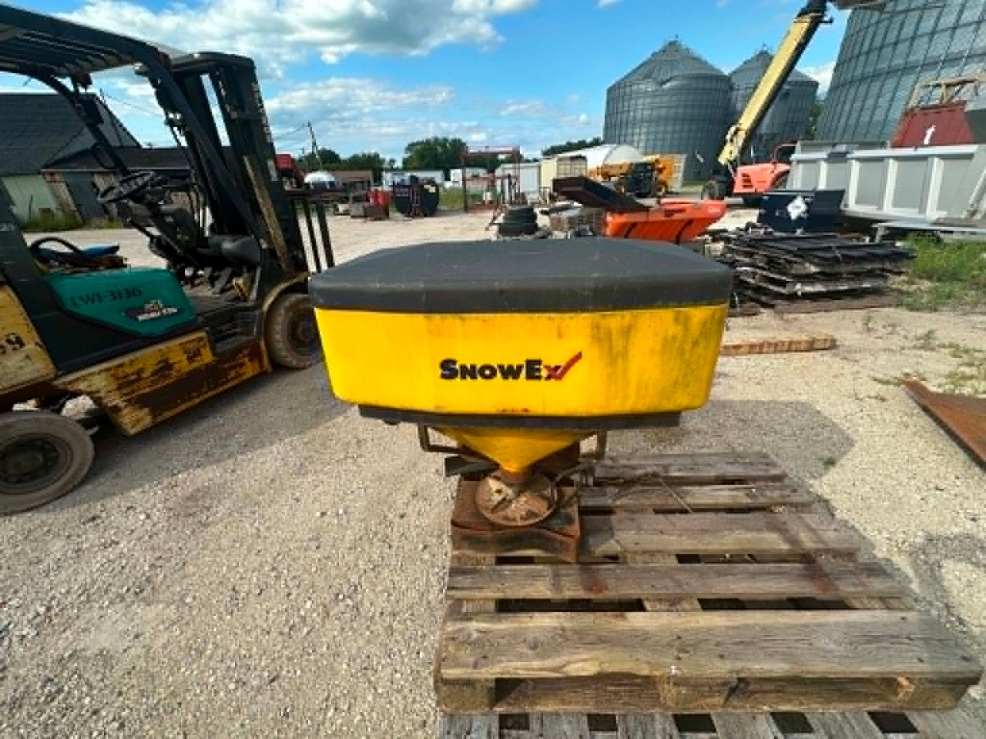 Yellow SnowEx spreader on a wooden pallet in a gravel equipment yard, with machinery and grain bins nearby