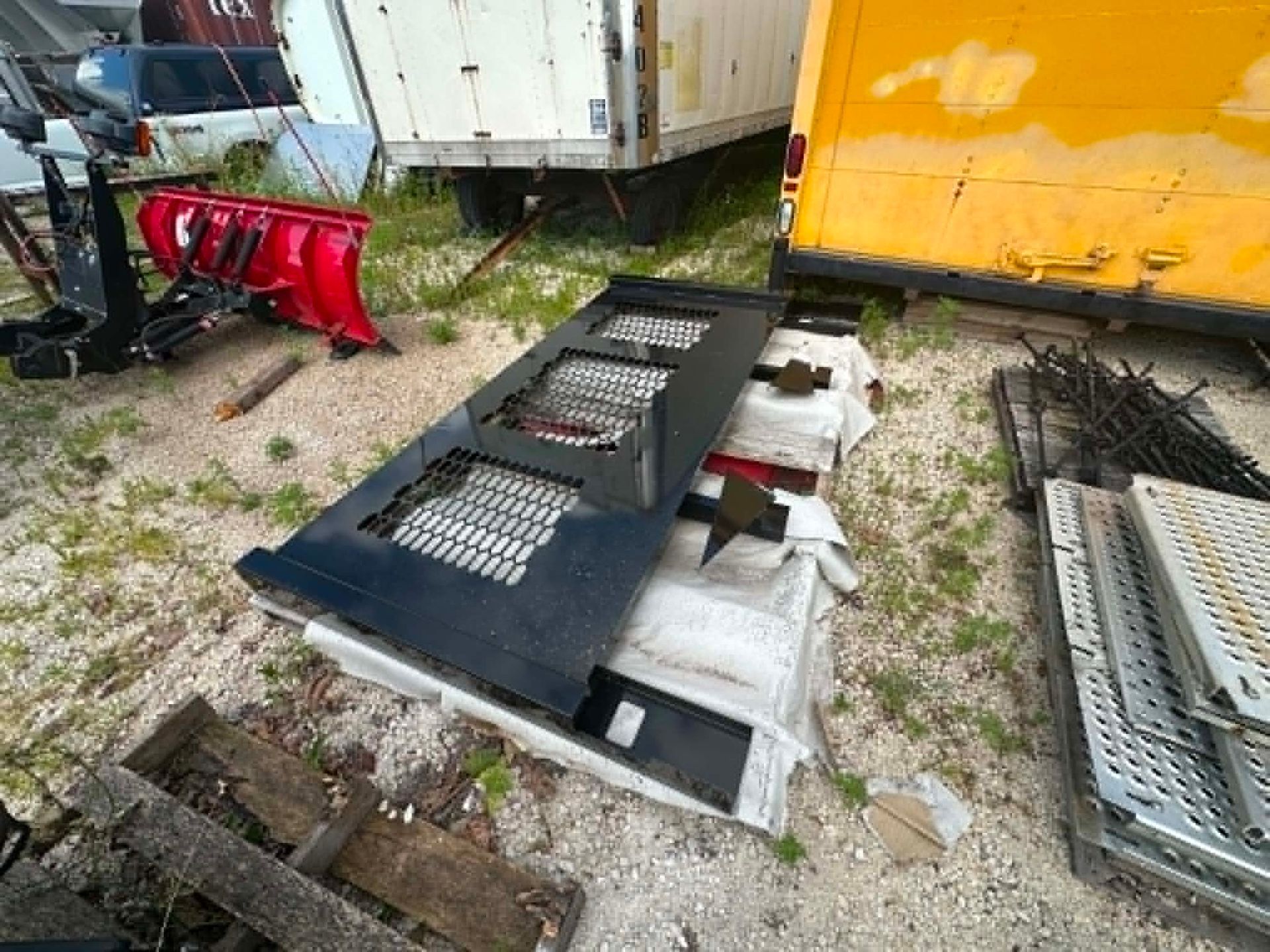 Black metal ramp or platform lying outdoors beside trailers and a yellow container, with scattered parts nearby