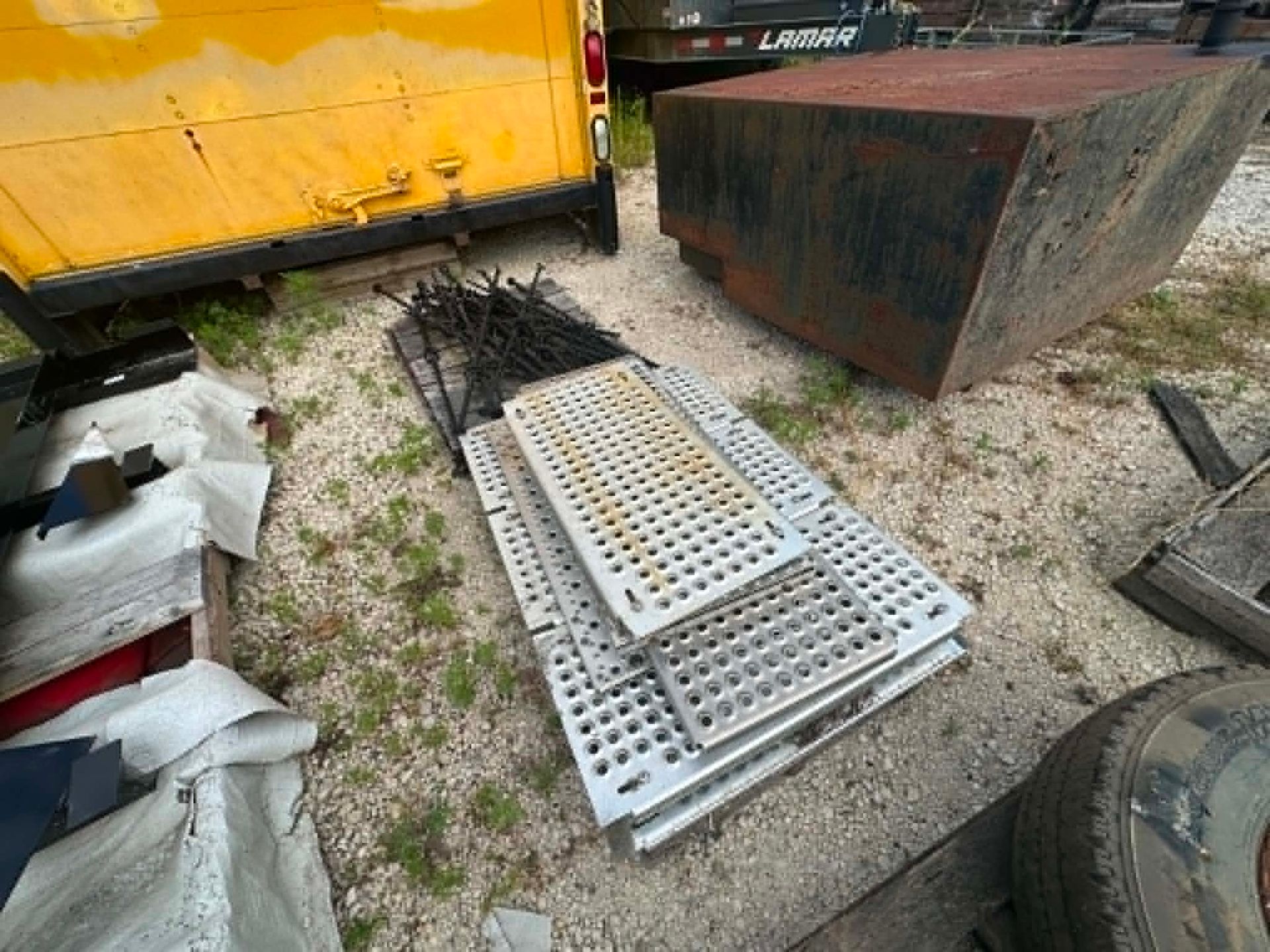 Metal grates and a black hose lying on gravel beside a yellow truck and rusted container