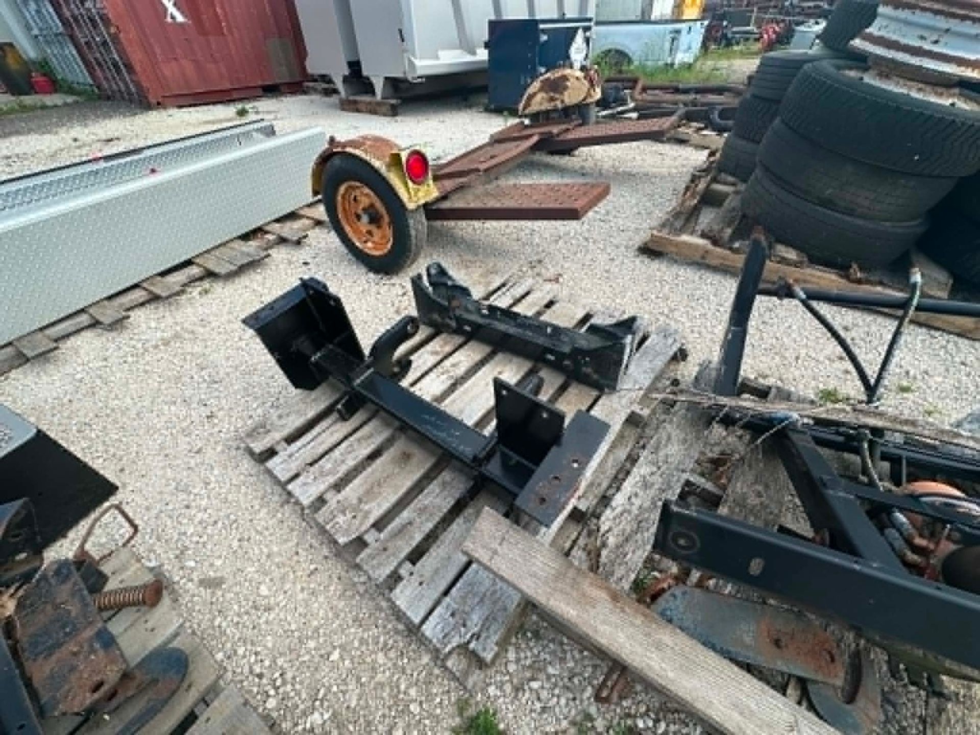 Scattered black metal parts and a wheel on wooden pallets in a gravel yard near stacked tires