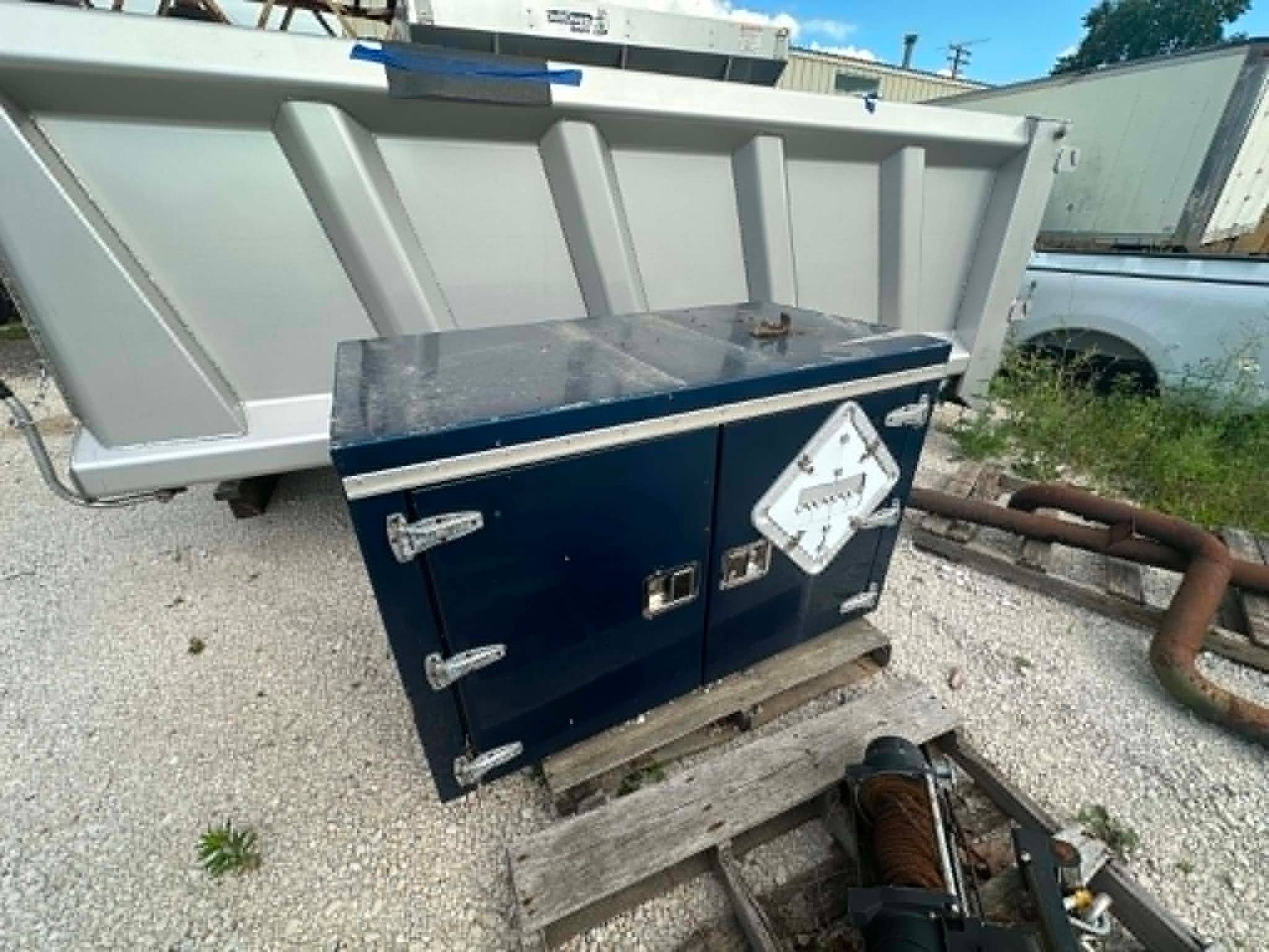 Blue outdoor generator beside a white dumpster in a yard, with pipes and debris nearby