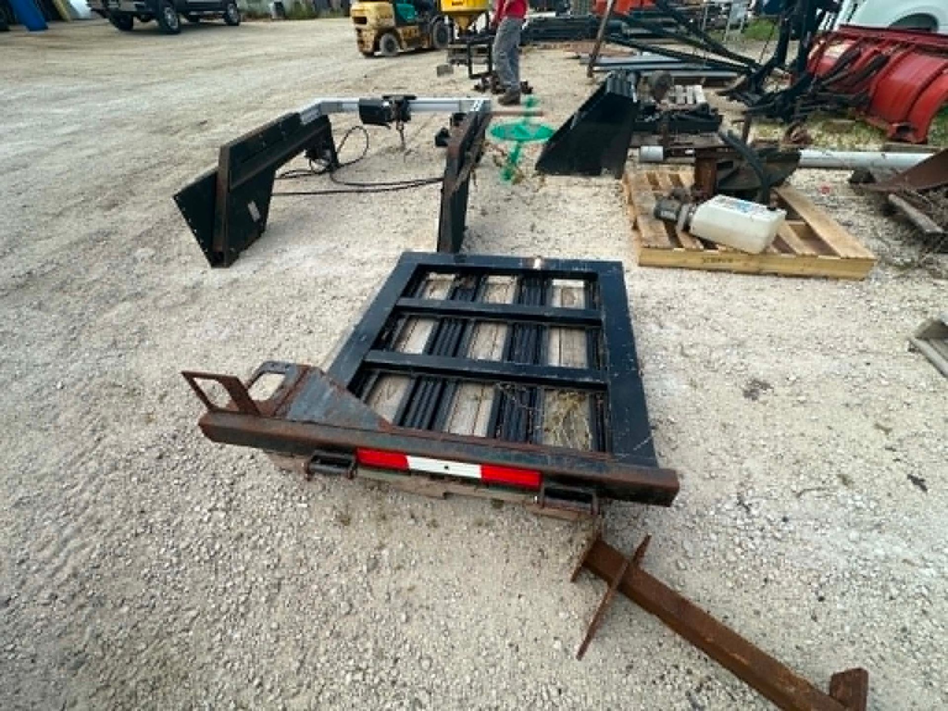 Flatbed trailer frame and black attachment parts in a gravel yard, with farm equipment in the background