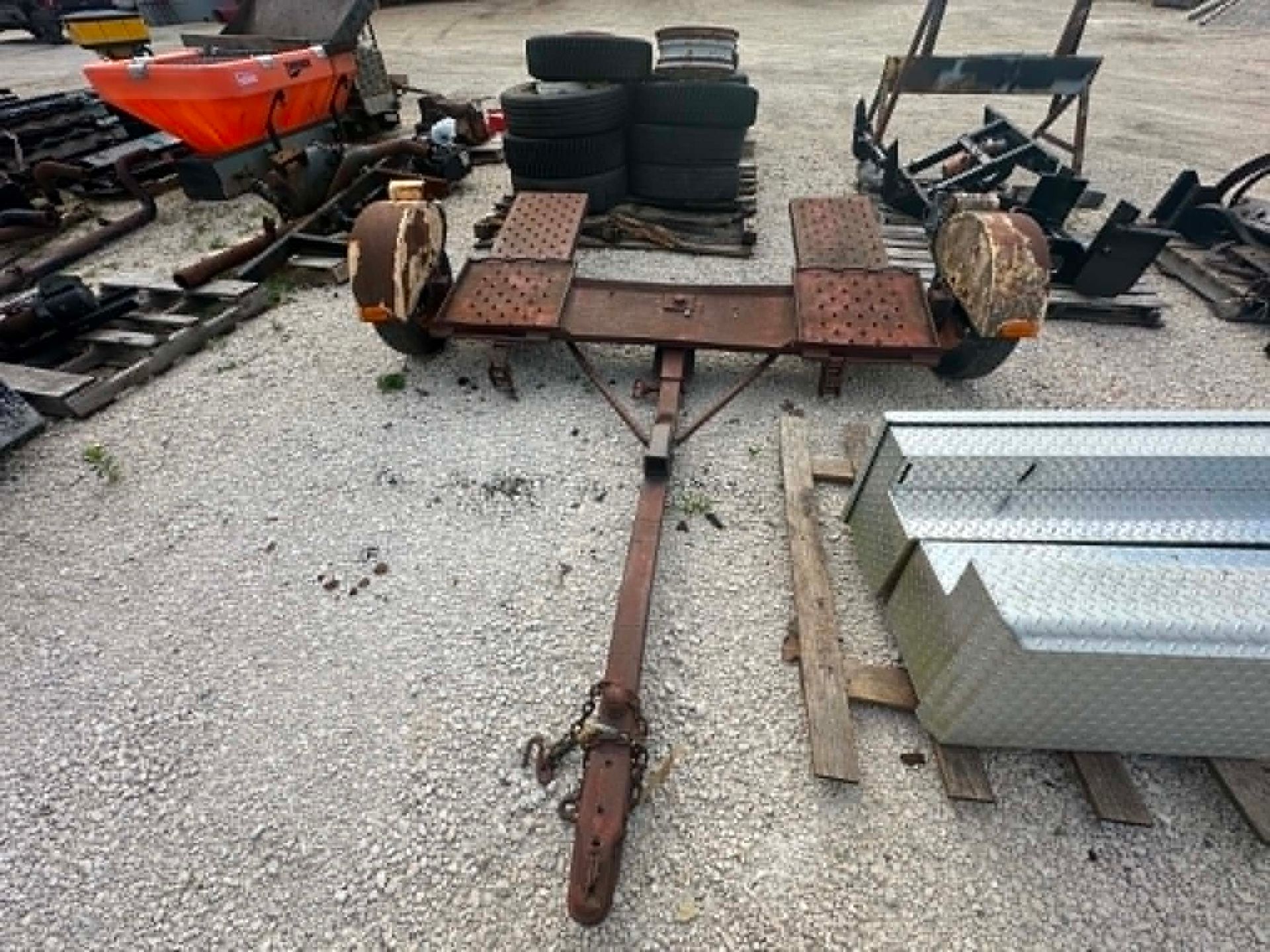 Rusty trailer frame with two wheels on a gravel lot, surrounded by tires and metal parts