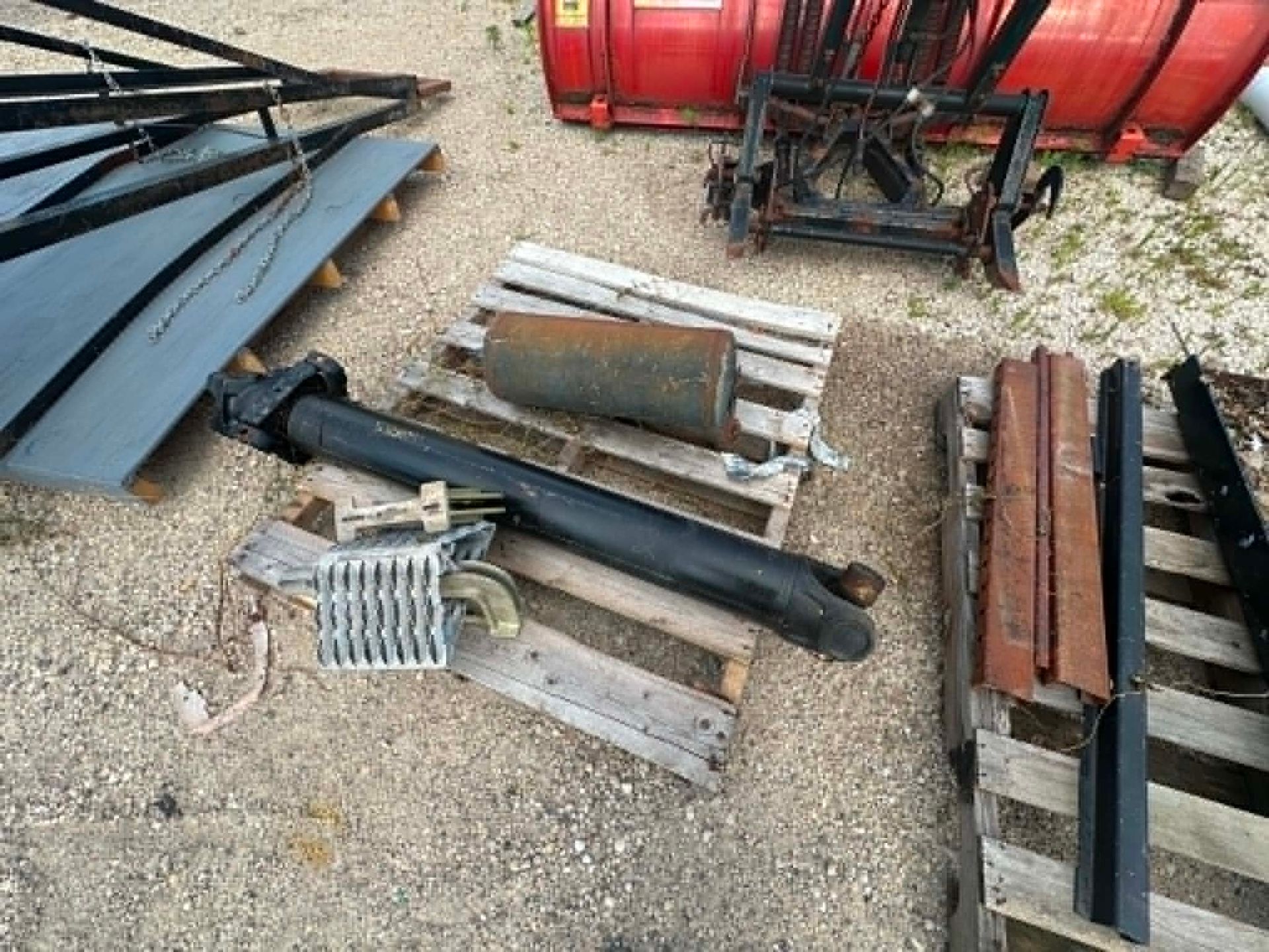 Assorted metal and farm equipment parts laid on gravel near red machinery and pallets