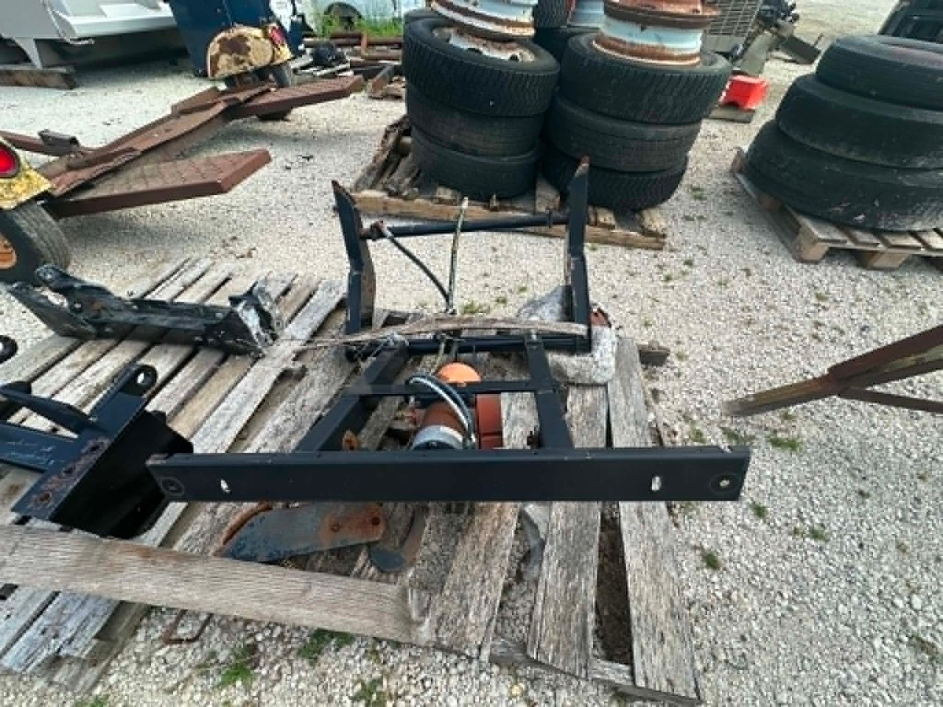 Rusty metal frame and axle assembly on a wooden pallet in a scrapyard, with stacked tires in the background