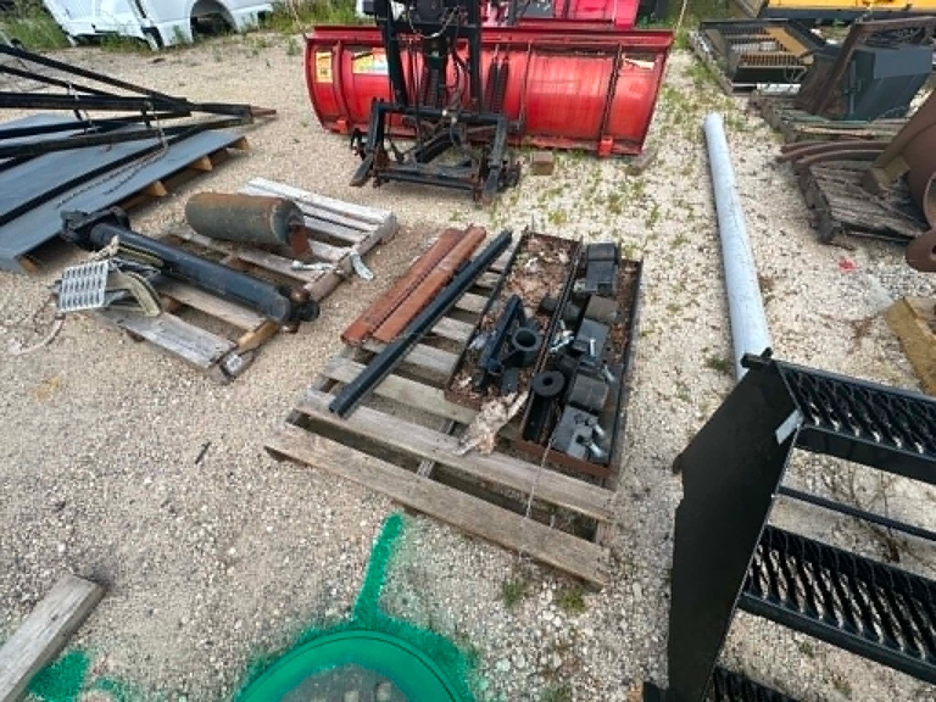 Outdoor yard with stacked metal farm equipment parts and a red snow plow attachment on gravel.