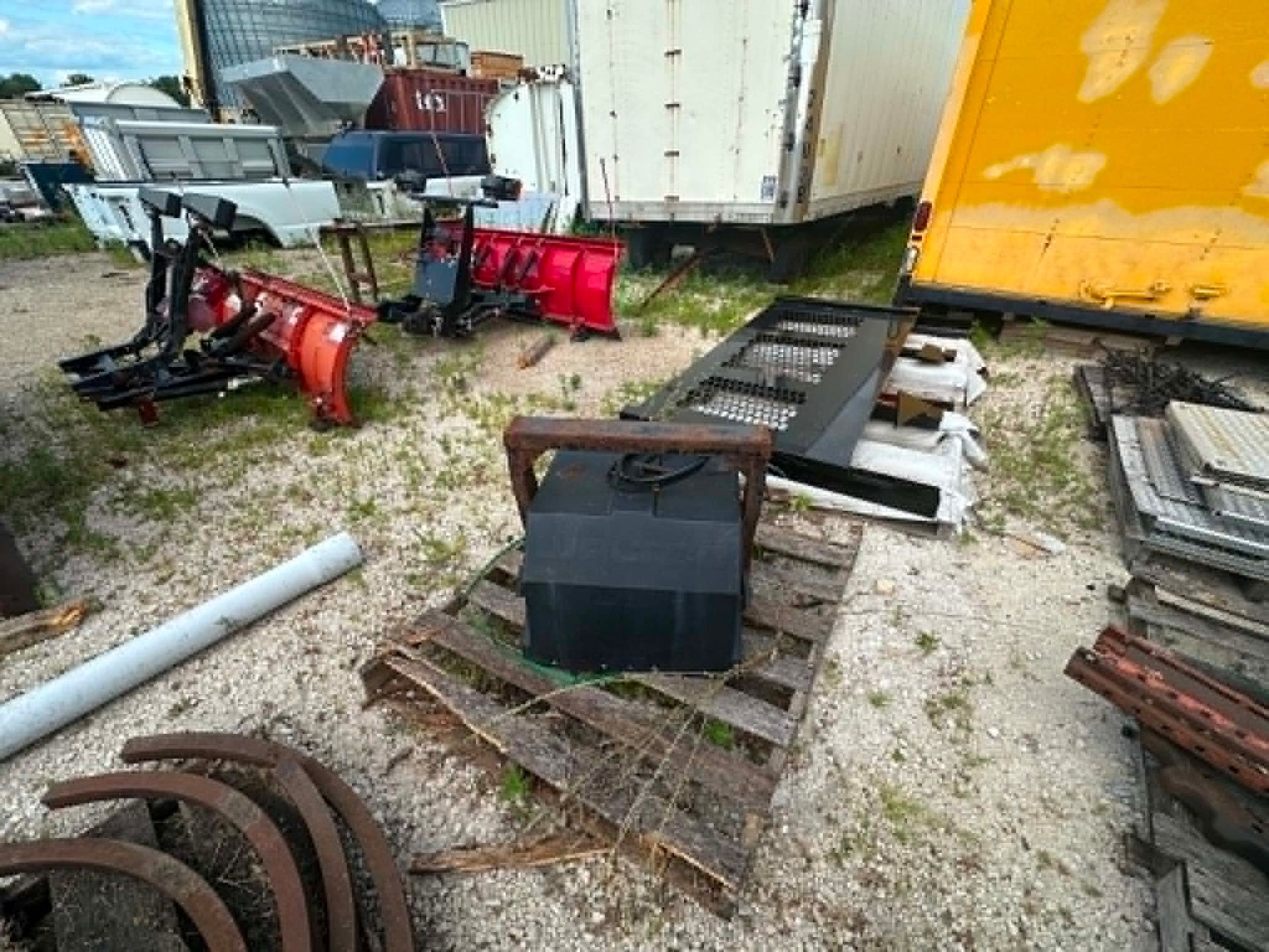Rusty farm equipment and scrap metal piled on gravel beside a yellow trailer outdoors