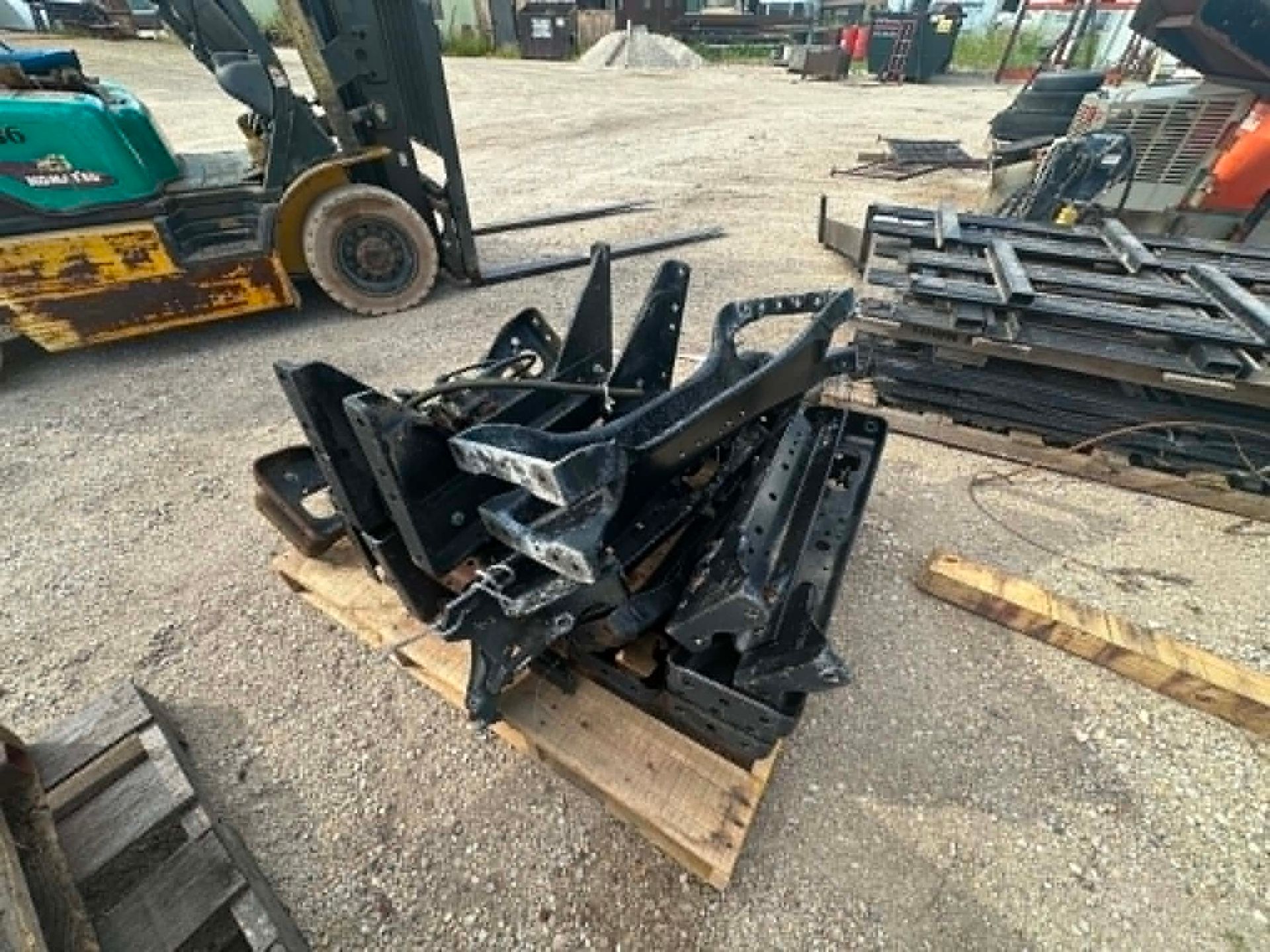 Stacked black metal equipment parts on wooden pallets in a scrapyard, with a forklift nearby.