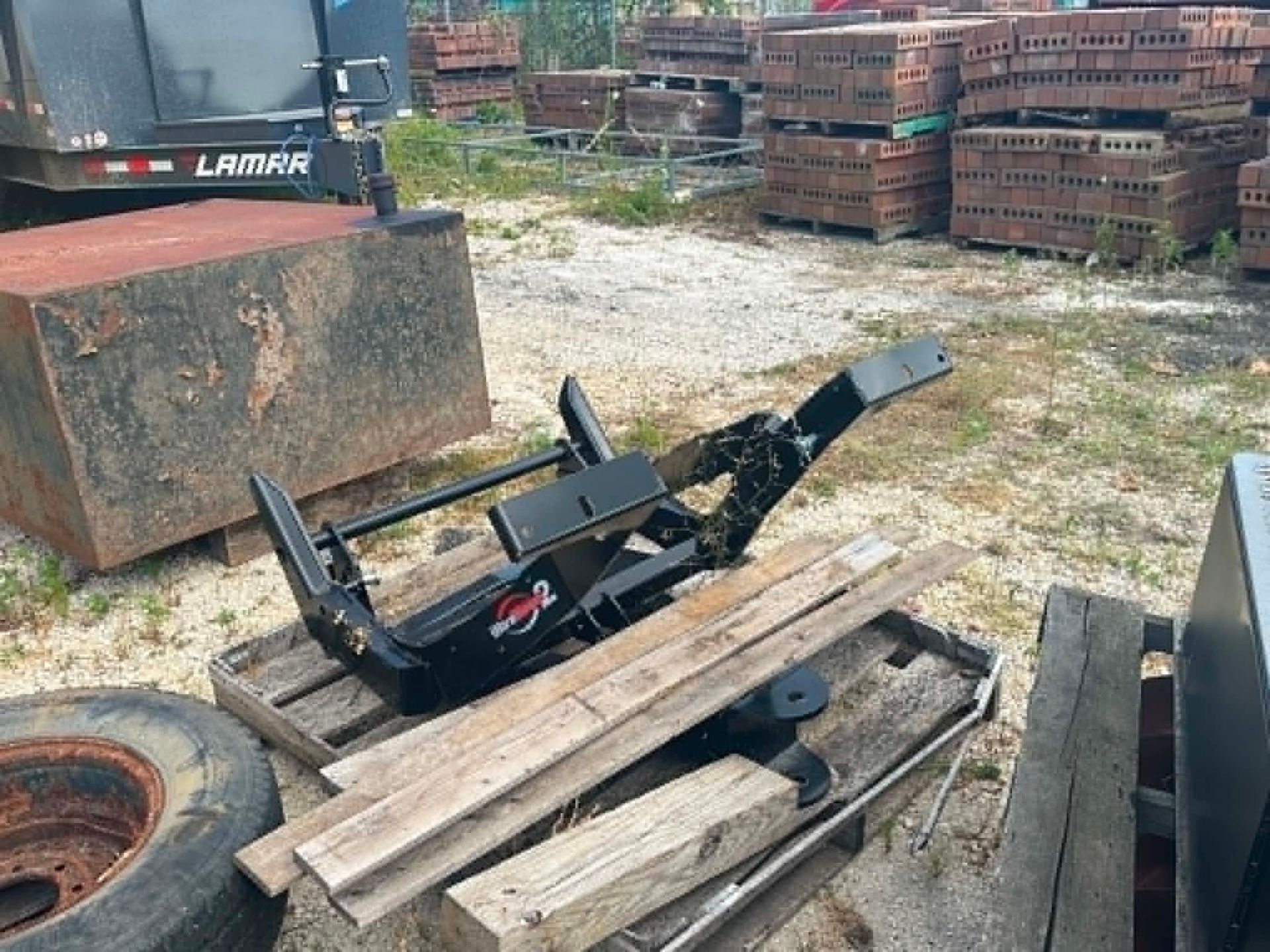 Forklift attachment sitting on wooden planks in a gravel yard, with stacked pallets in the background.