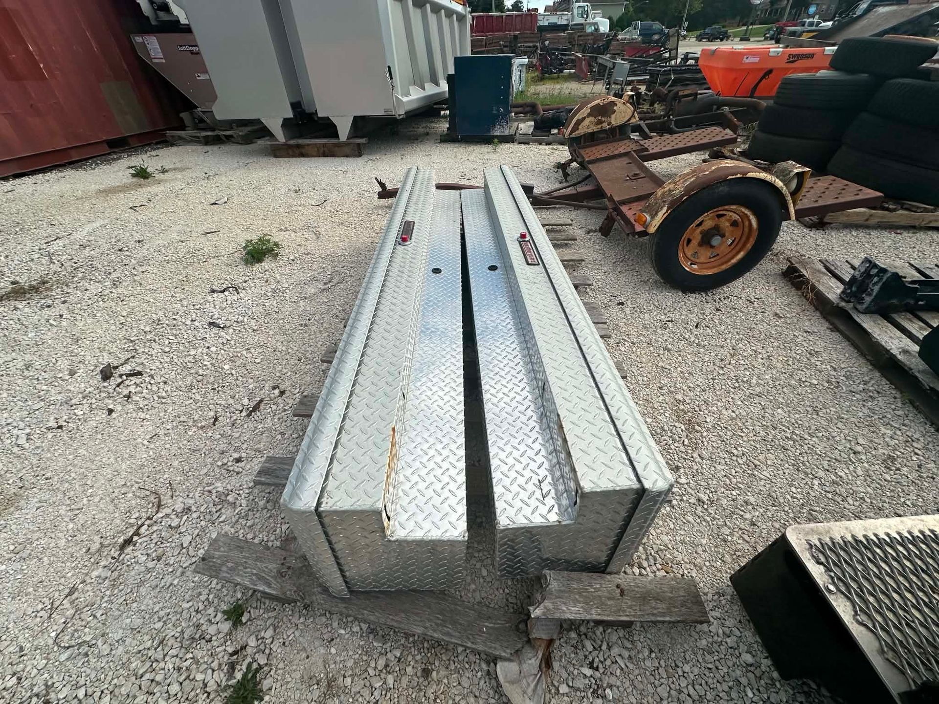 Long silver metal beams stacked on a gravel lot beside equipment and scrap.