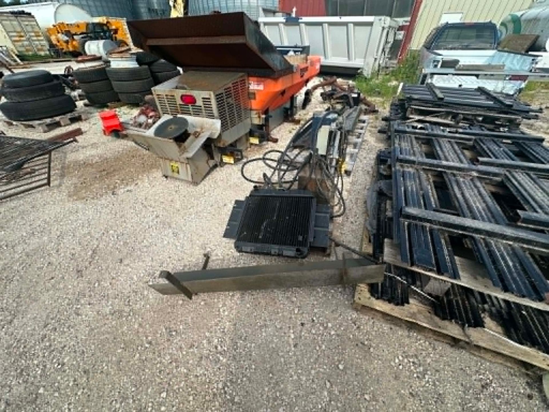 Cluttered construction yard with stacked metal parts, tires, tools, and equipment on gravel.