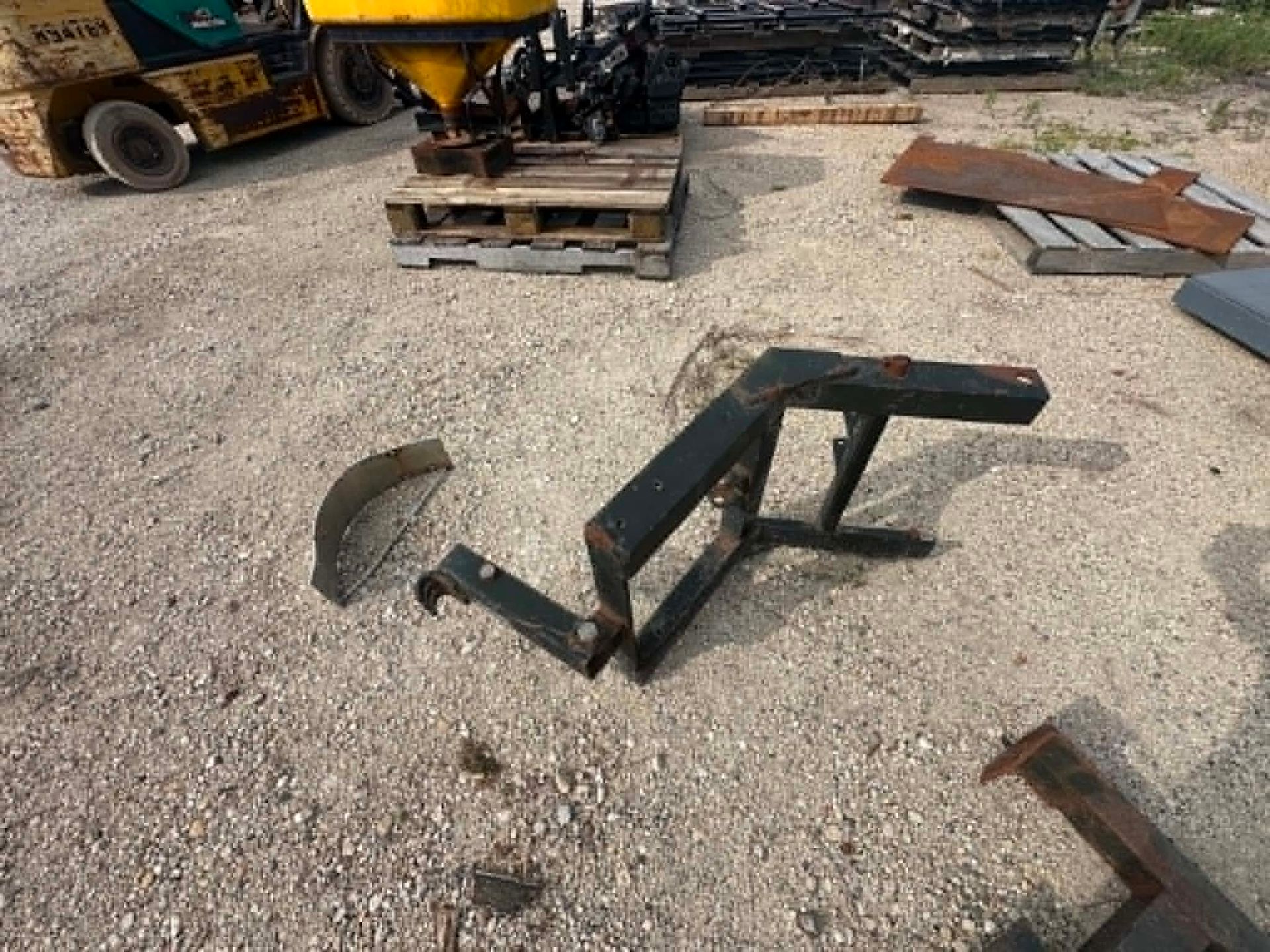 Bent metal farm implement parts on gravel in a work yard, with pallets and equipment in the background