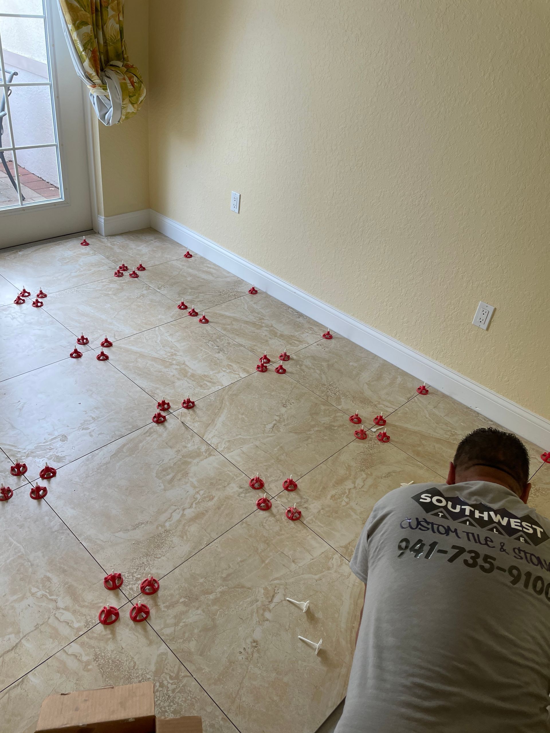 Person kneeling on tile floor beside newly installed carpet tiles, with red spacers scattered across the room.