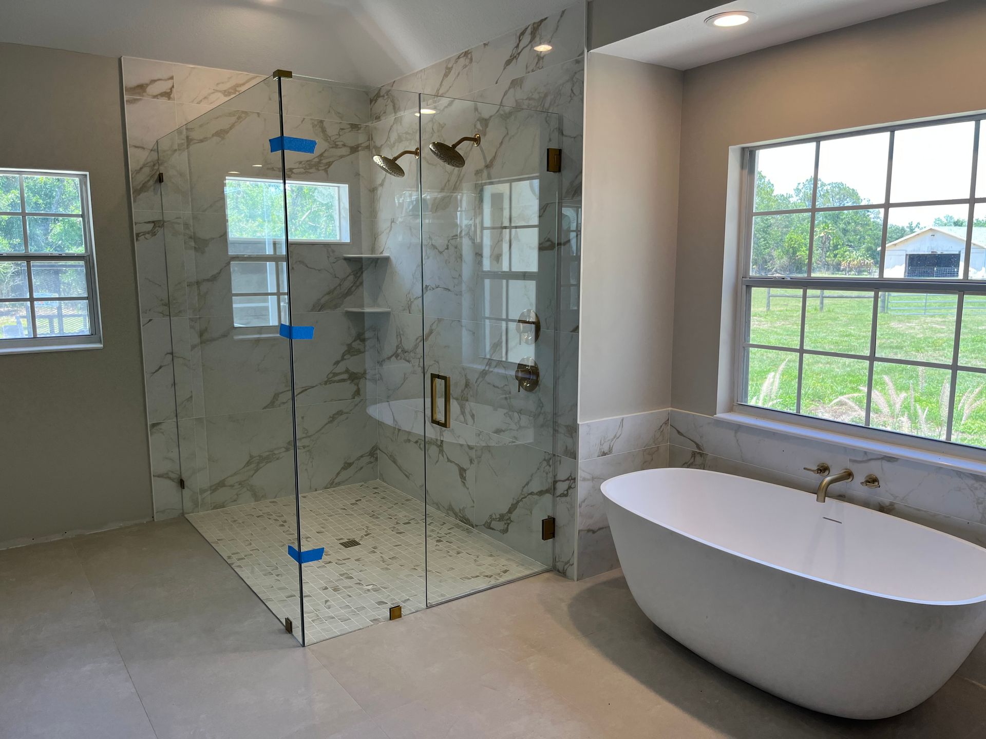 Modern bathroom with glass shower, marble tile, and freestanding white bathtub by a window