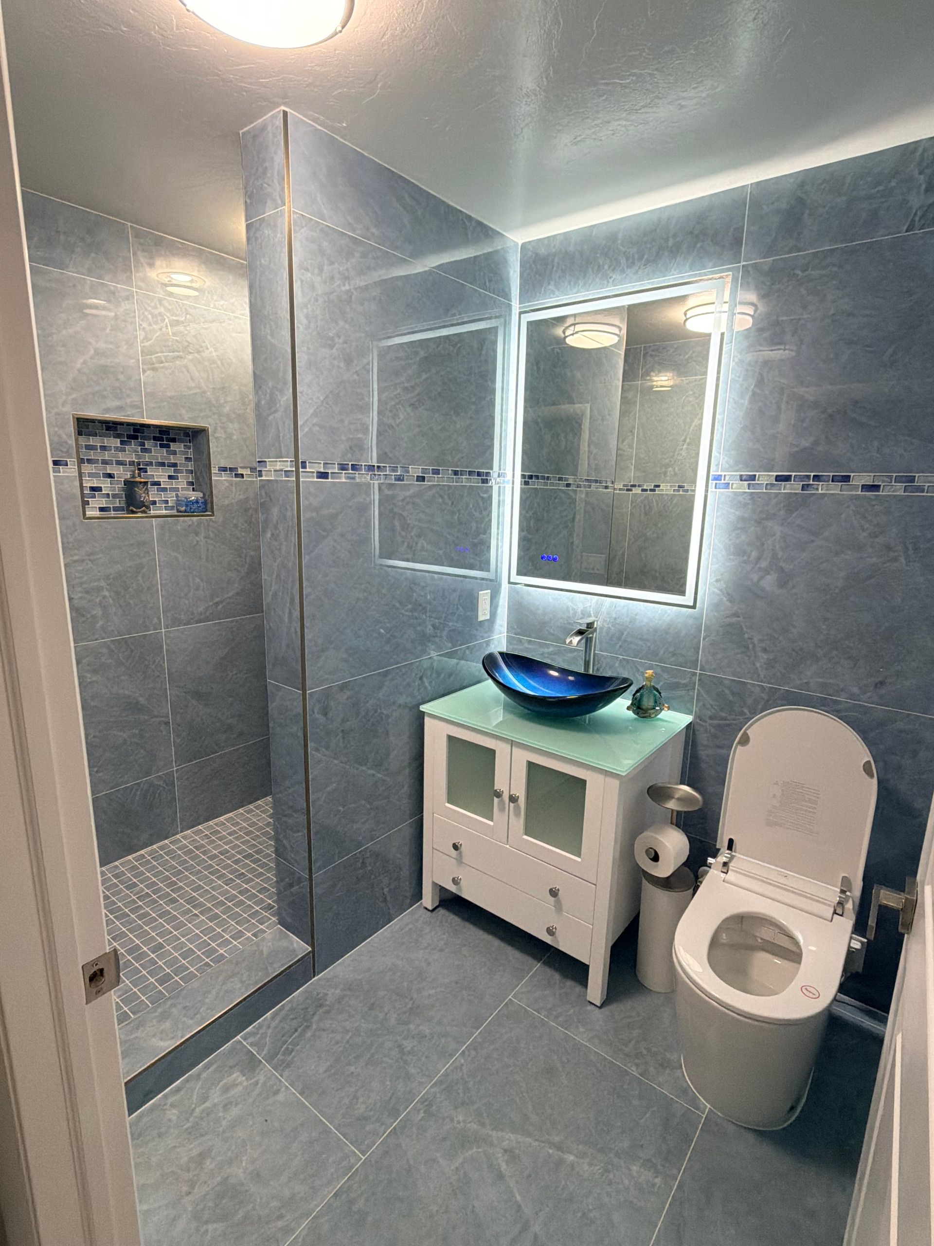 Bright blue-gray tiled bathroom with shower, mirror sink vanity, and toilet under wall light.