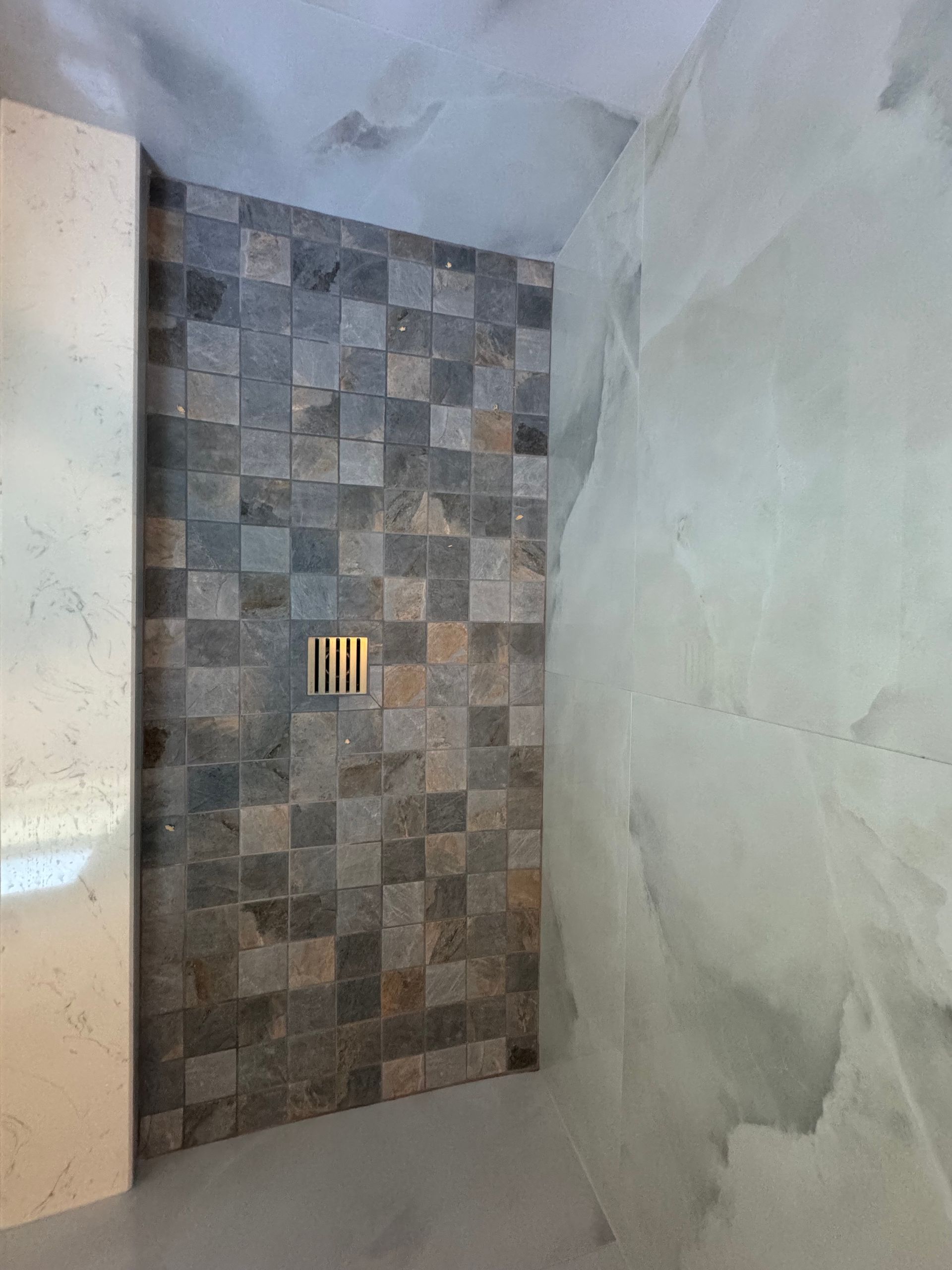 Small tiled shower alcove with gray mosaic wall and a gold drain fixture, surrounded by unfinished plaster walls