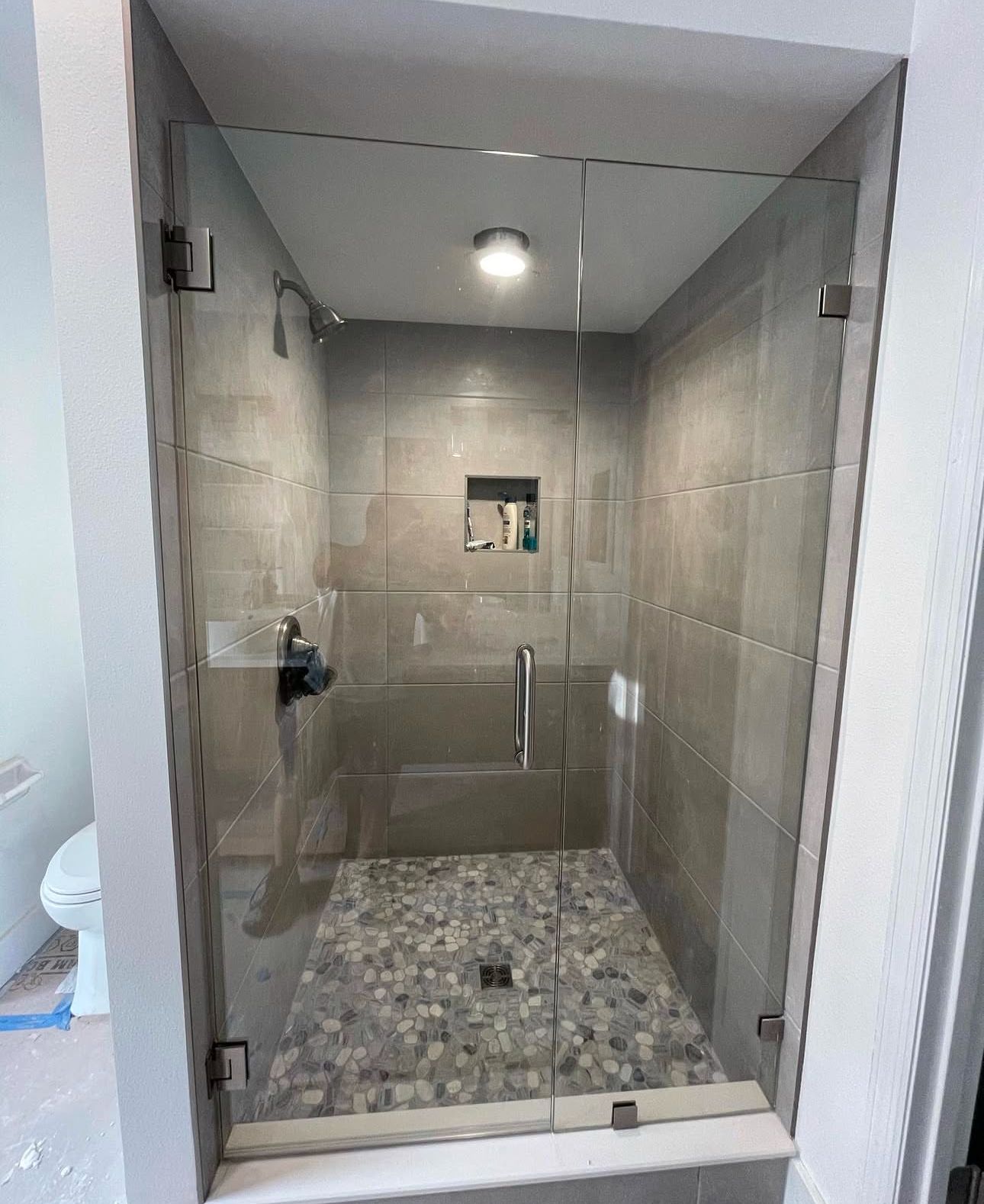 Glass shower stall with gray tile walls and pebble floor, viewed from the bathroom entry.