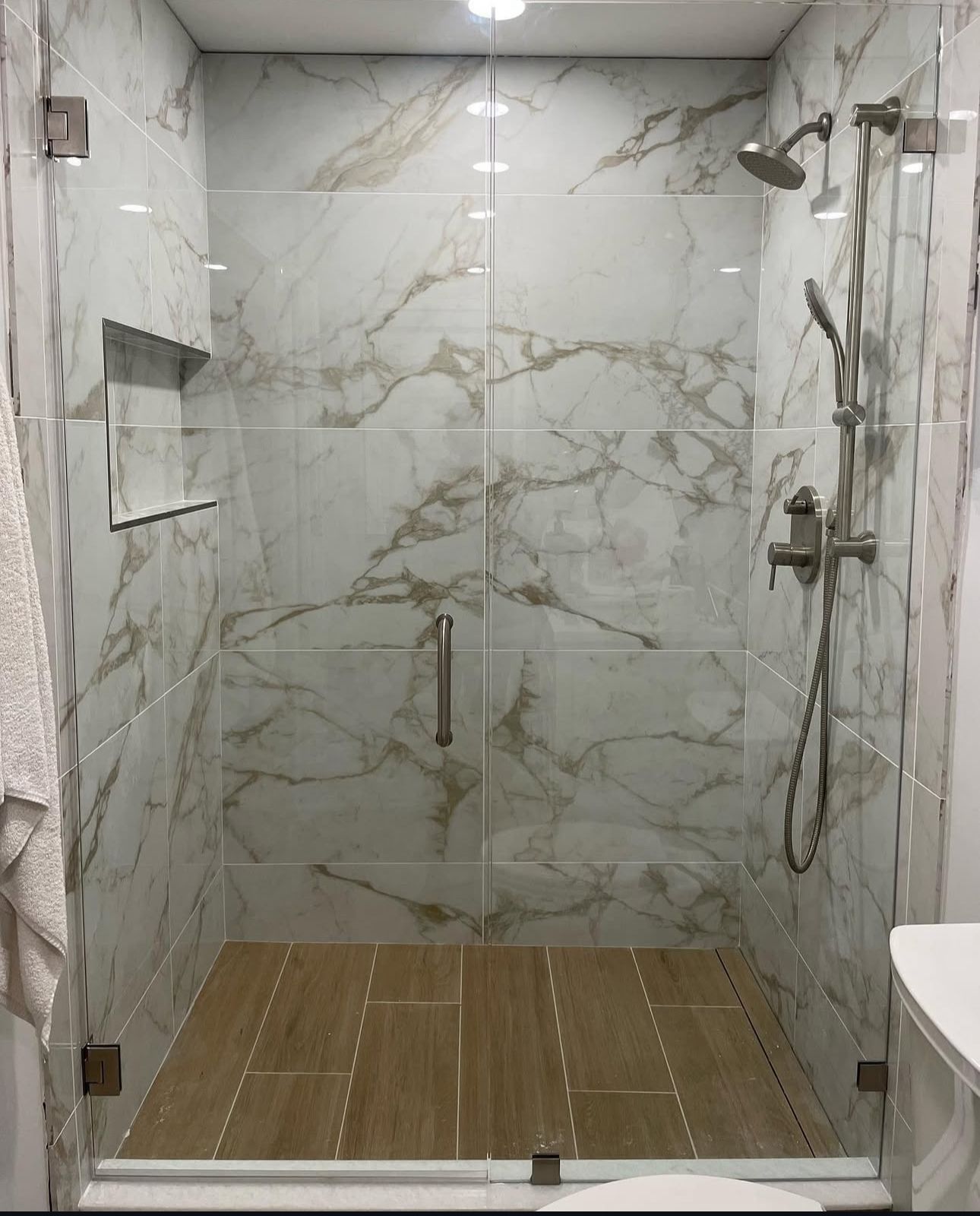 Modern tiled walk-in shower with glass doors, rain showerhead, handheld sprayer, and bamboo mat floor mat