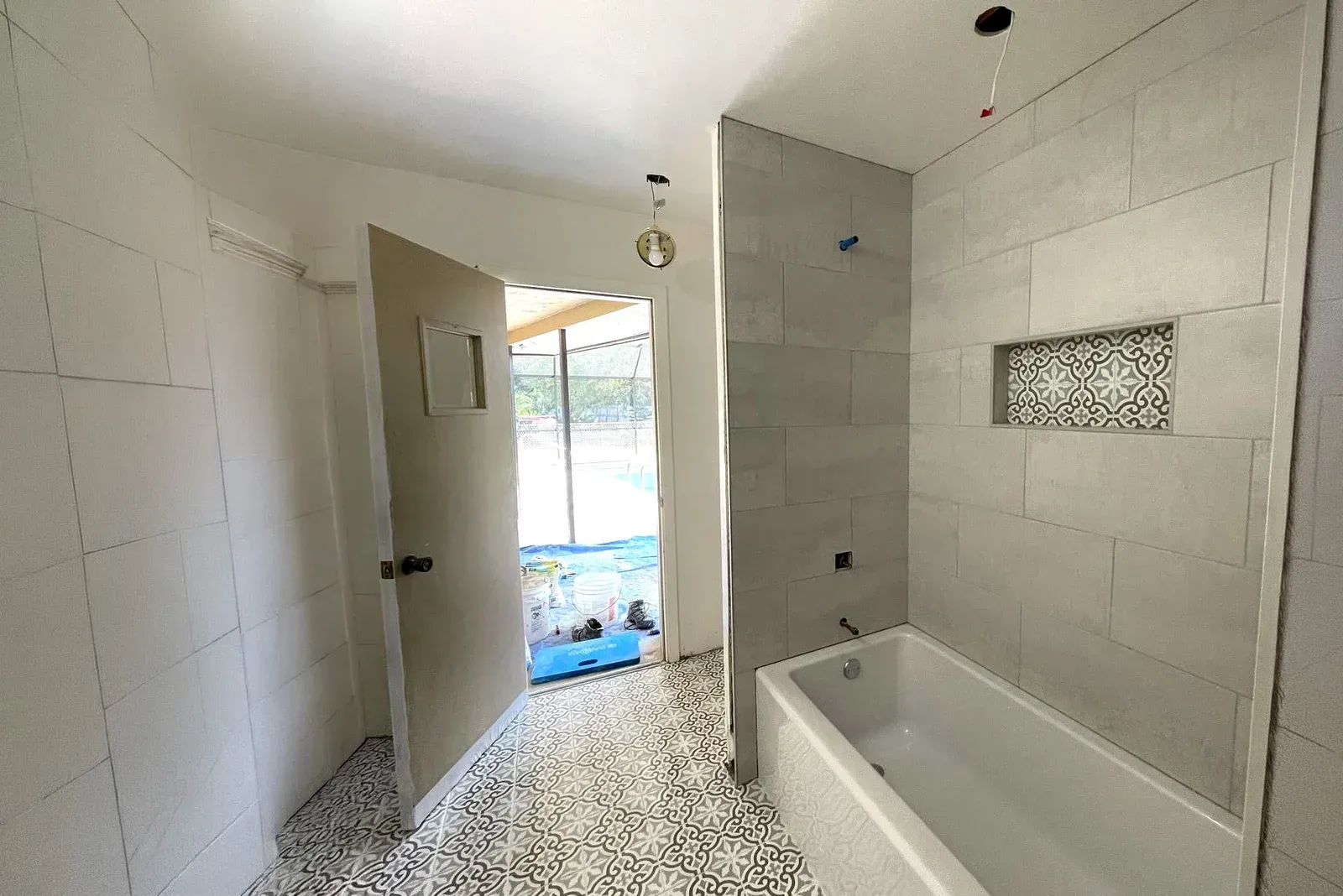 Bright bathroom with white tiled walls, a bathtub, and an open door to the outside.