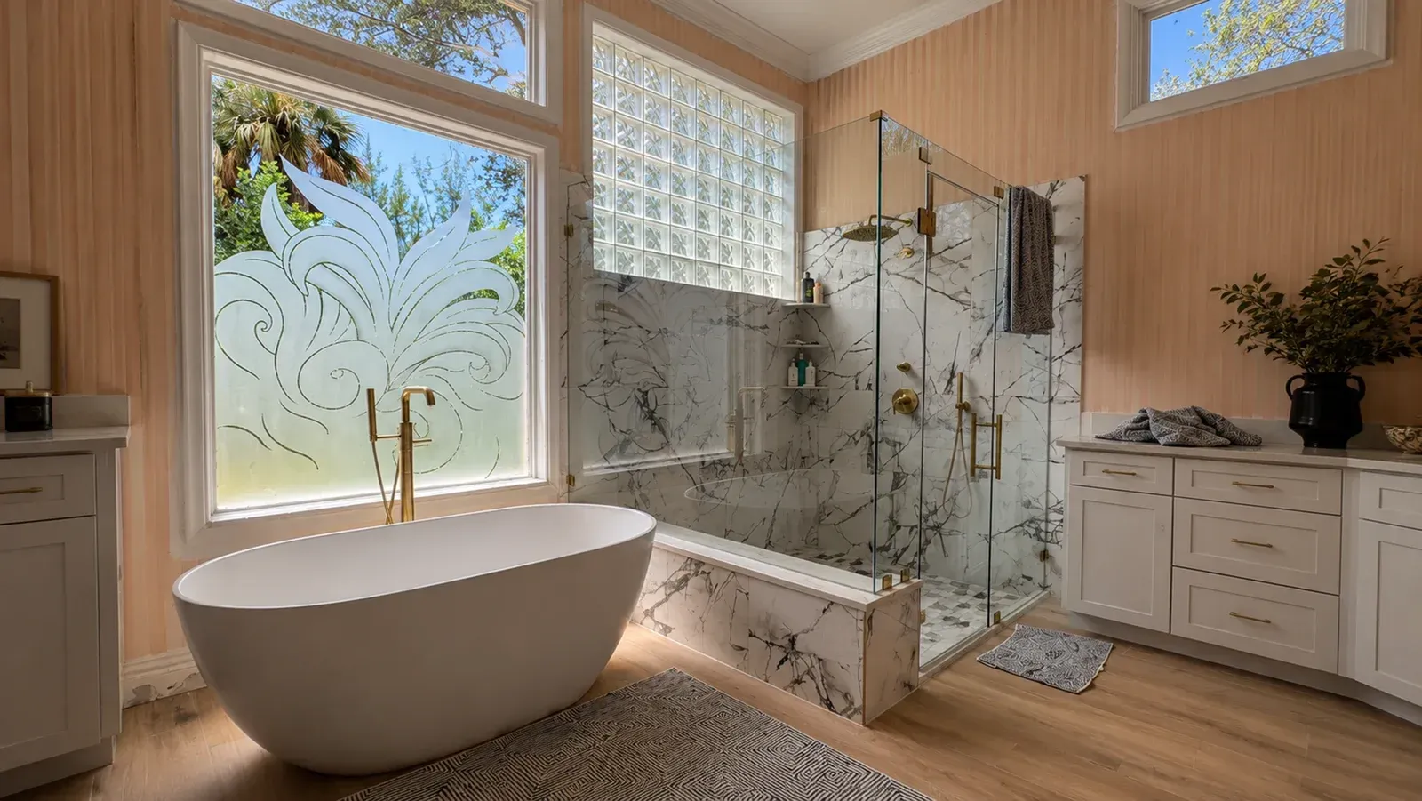Bright luxury bathroom with freestanding tub, glass shower, and white vanities by large windows.