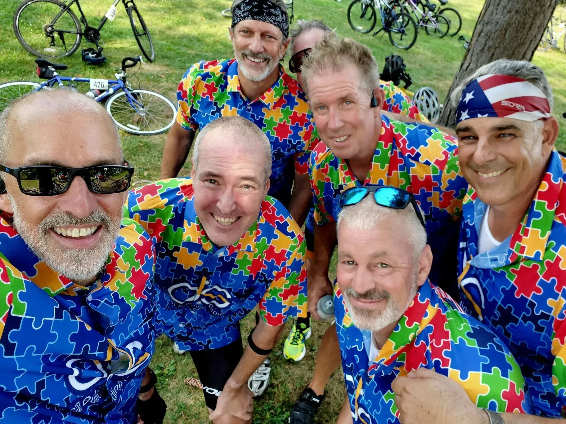 Group of men in colorful puzzle-piece shirts smiling, posing with bikes, outdoors on a sunny day.