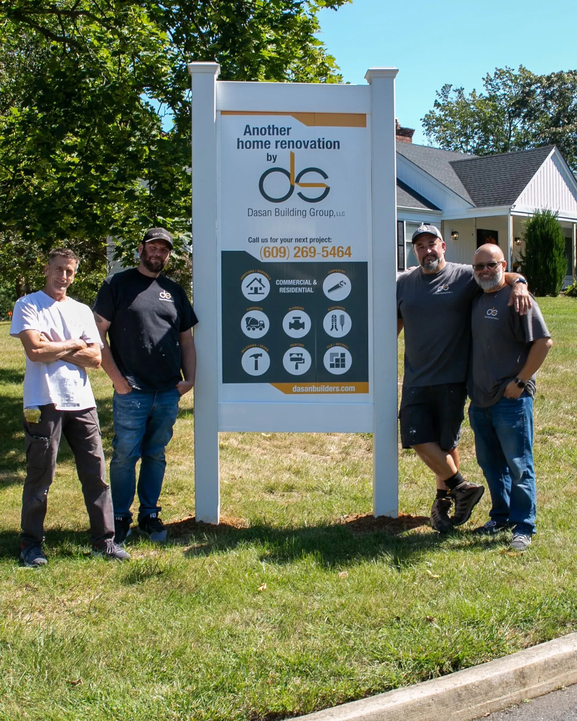 Four men stand by a sign with a building in the background. The sign is white with business information.