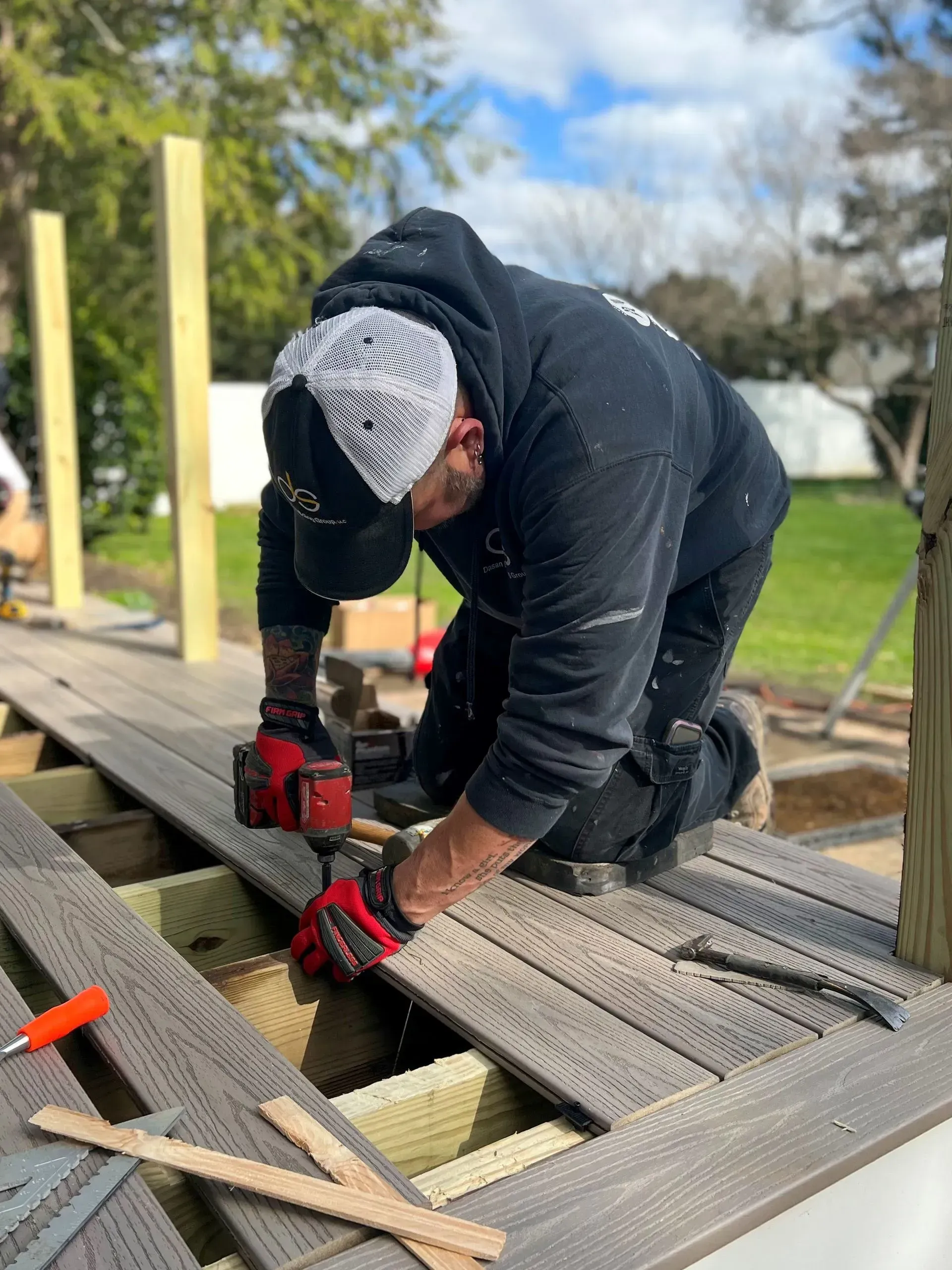 Construction worker kneels on a deck, using a drill to secure boards. He wears a black hoodie, hat, and work gloves.