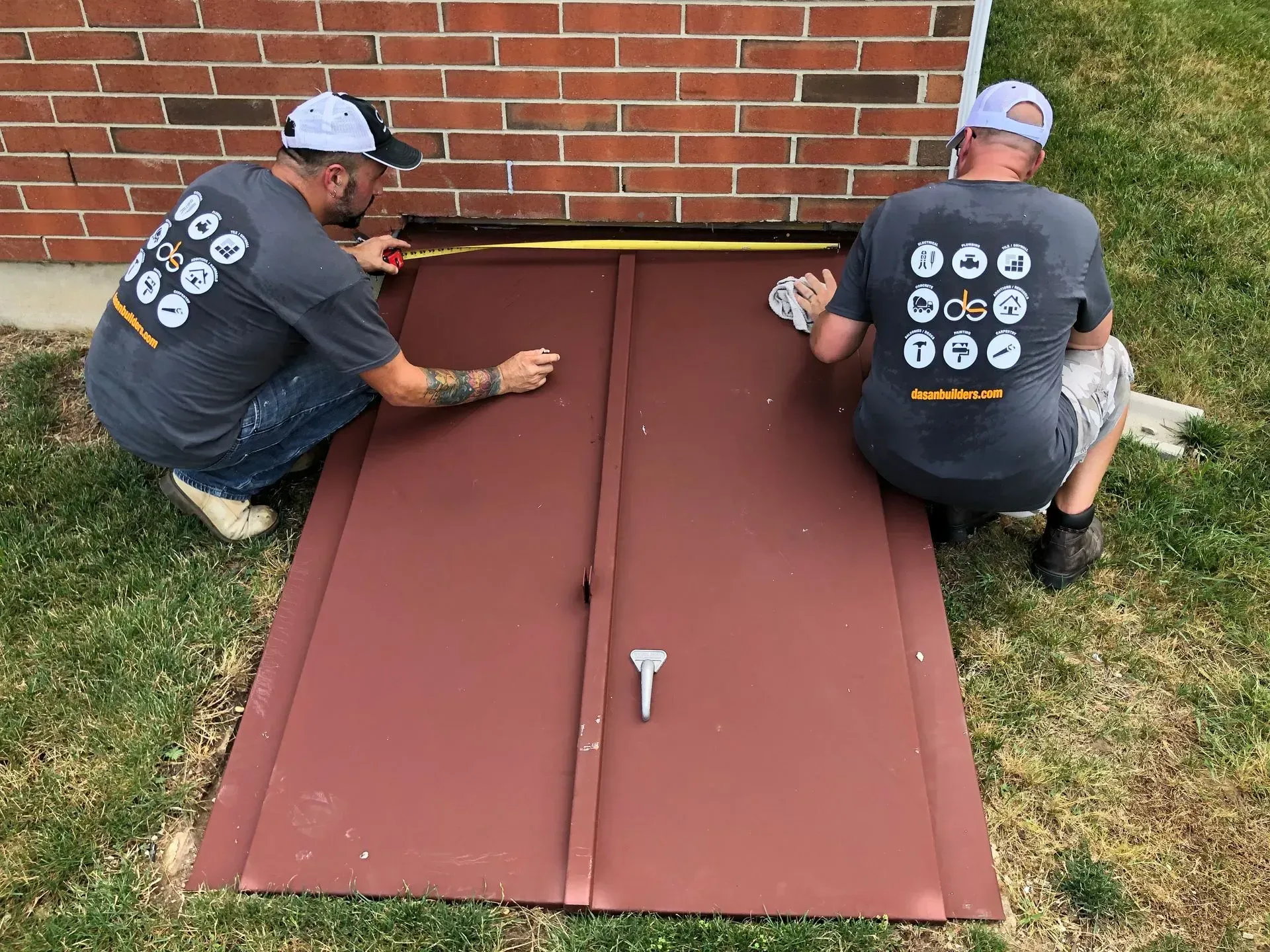 Two men painting a brown basement door outdoors, against a brick wall and grass.