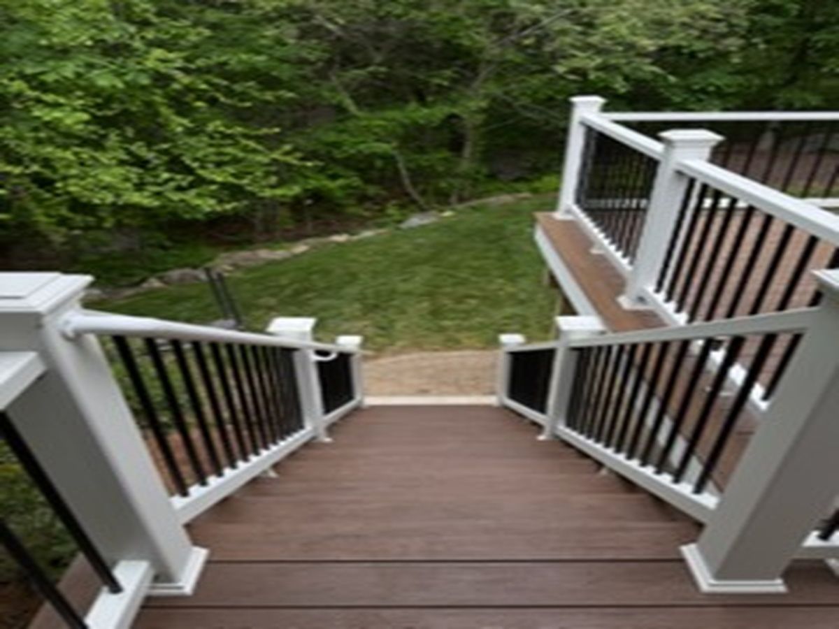 Wooden deck stairs descending to a grassy yard, bordered by white railings and surrounded by green trees.