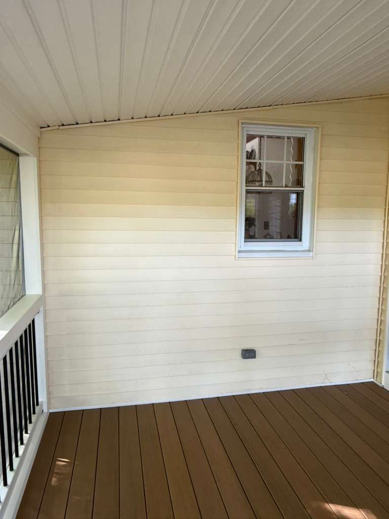 Covered porch with white siding, brown deck floor, and a small window on the wall.