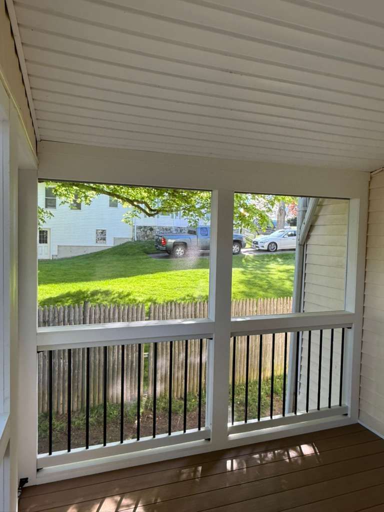 Covered porch overlooking a sunny front yard, street, and parked cars through white railing and columns