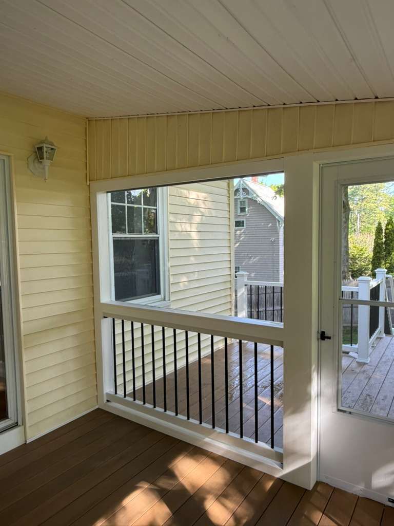 Covered porch with beige siding, brown floor, white railing, and an open door to a sunny deck.