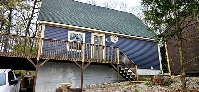 Blue house with deck and stairs on a wooded hillside, with a car partly visible at left.