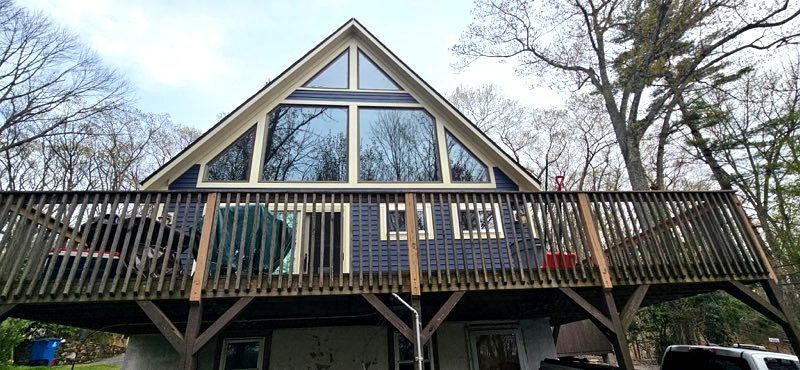 A-frame cabin deck with dark blue siding and tall windows, viewed from below among bare trees