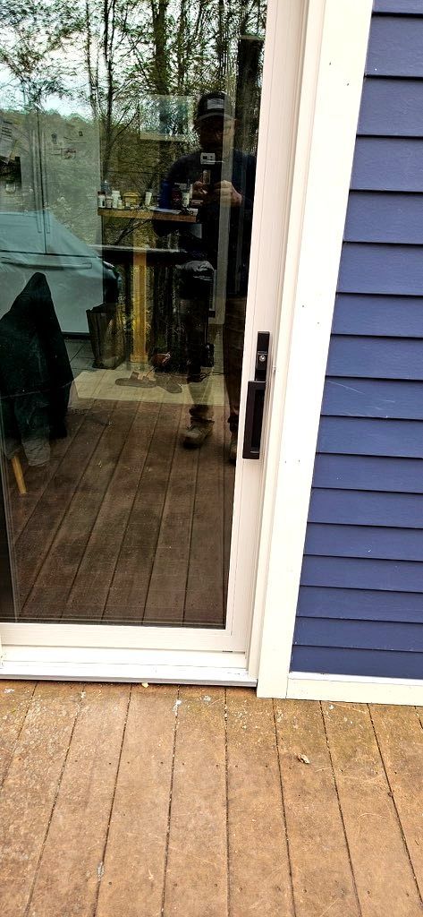 Glass patio door beside blue siding and a wooden deck, with a reflection of a person indoors.