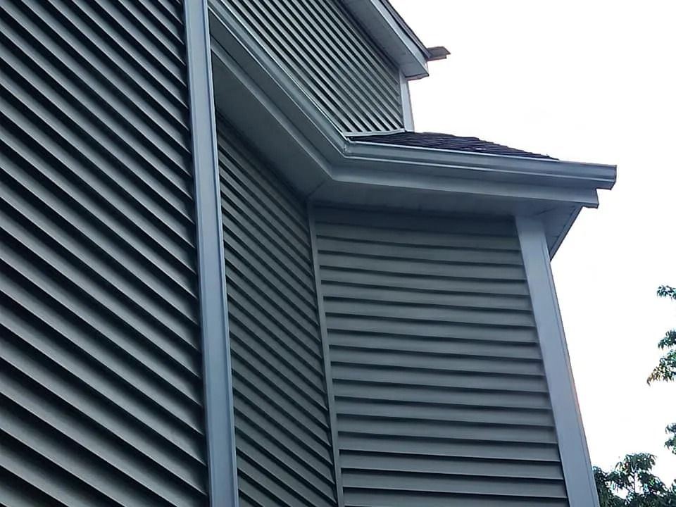 Corner of a house with green horizontal siding, white trim, and a gutter against a cloudy sky.