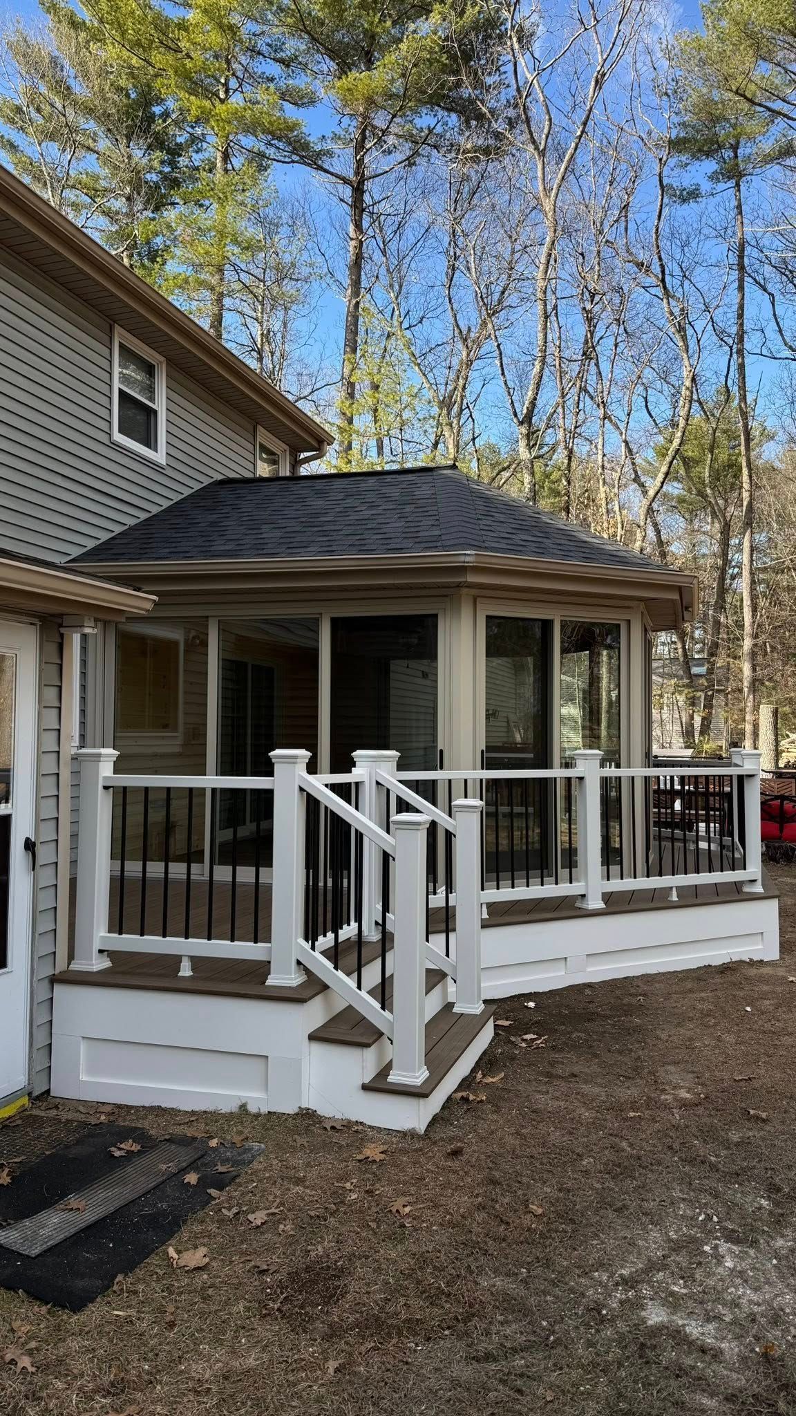 A tan-sided house with a screen porch and white deck featuring black balusters, set against a backdrop of bare trees.