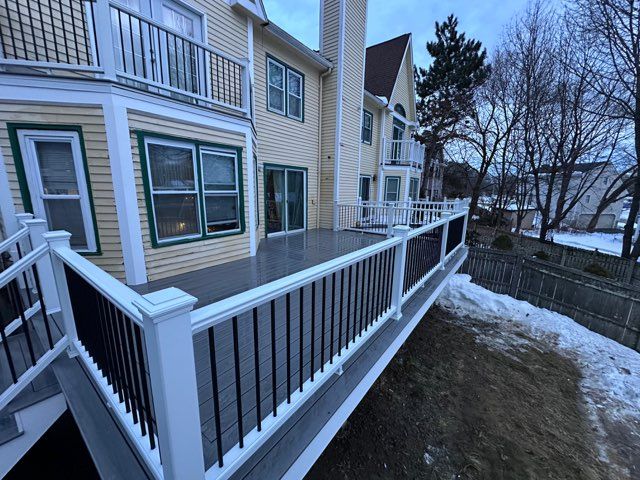 A residential deck with grey composite planks, white railings, and black balusters attached to a yellow house in winter.