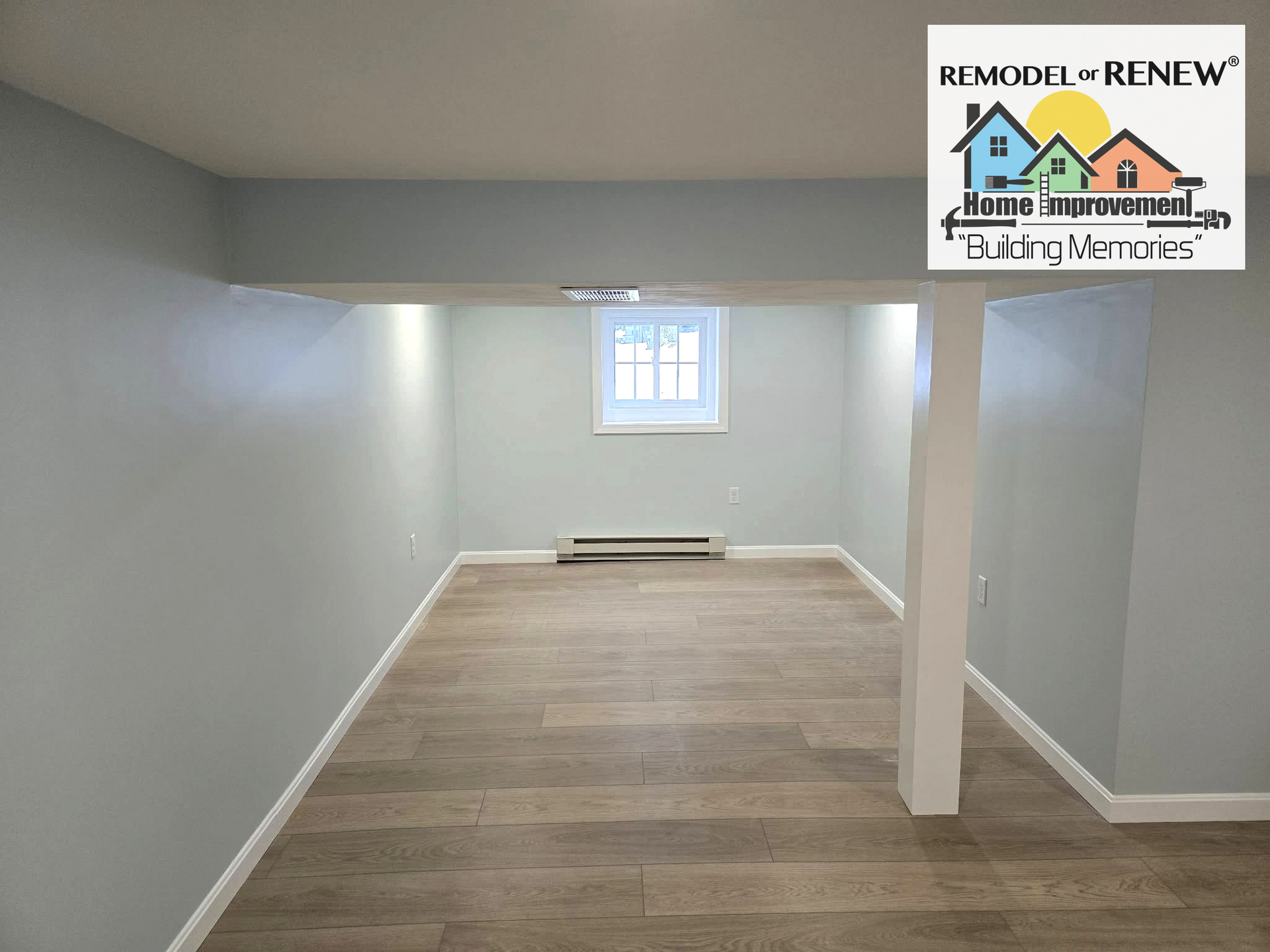 A light-blue finished basement room with wood-look flooring, a window, baseboard heating, and a structural post.