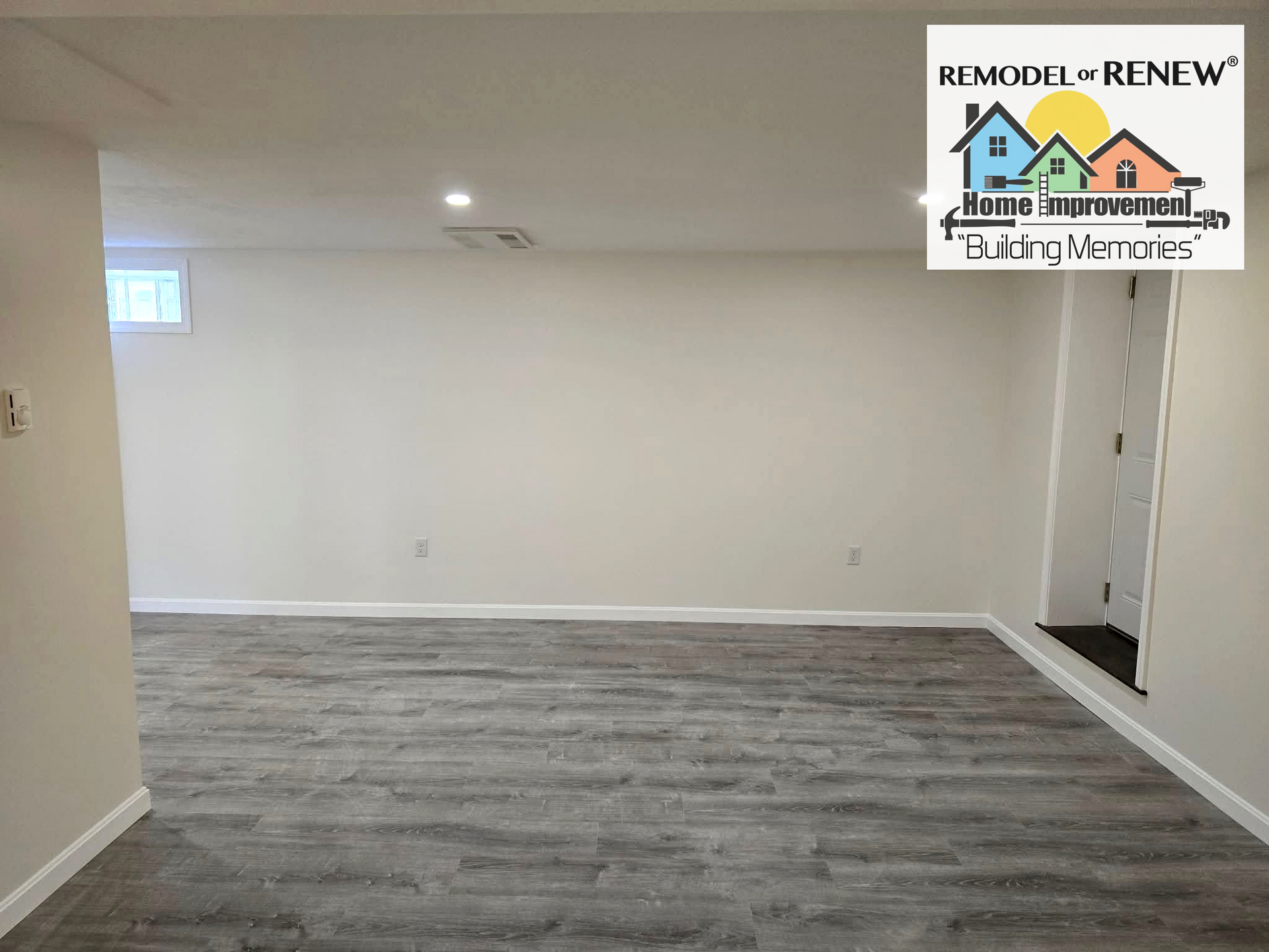 A basement interior with light-colored walls, recessed lighting, gray wood-look floors, a small window, and a doorway.