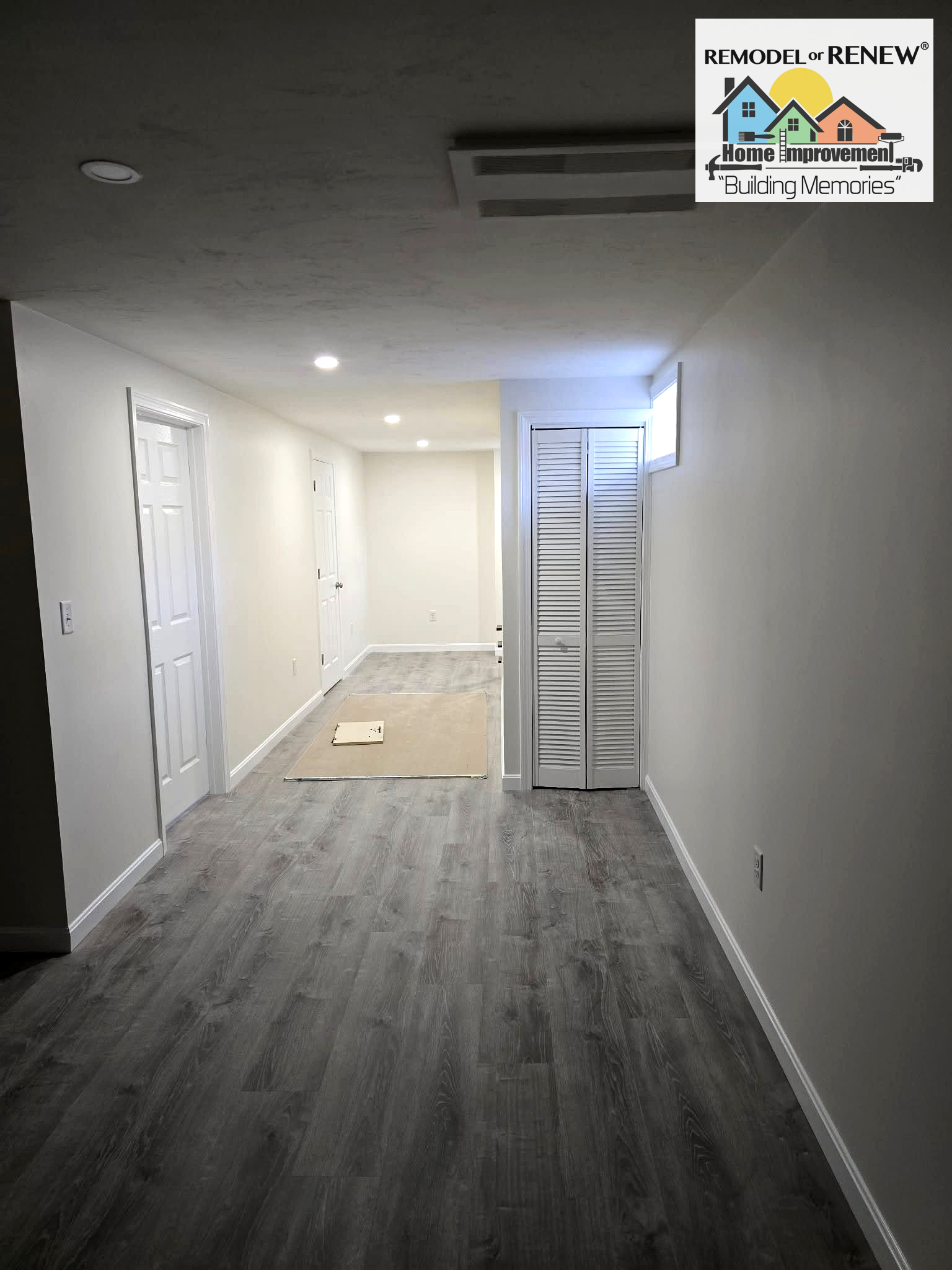 A basement hallway with gray wood-look flooring, white walls, two white interior doors, and a set of bifold closet doors.