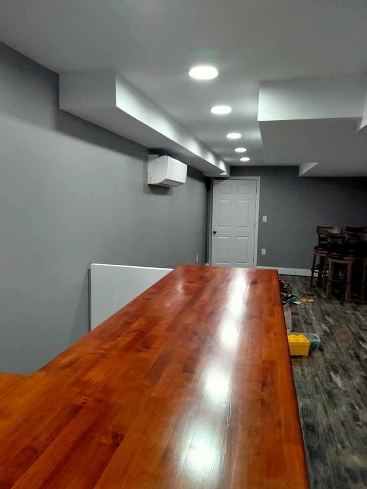 A wooden bar in a basement with gray walls, white ceiling beams, and recessed lights.