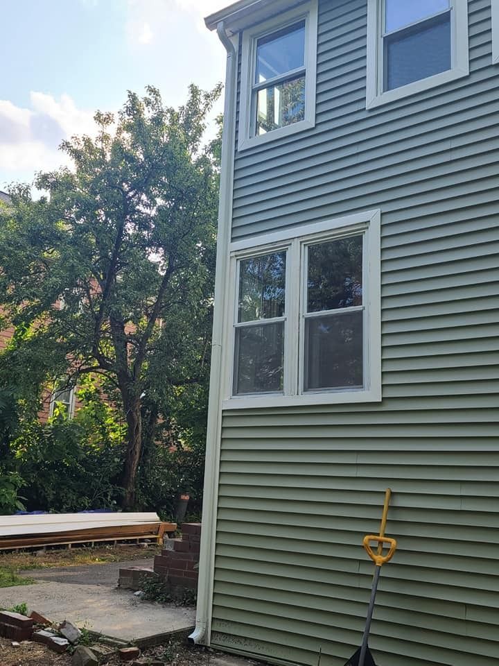 Green-sided house with white window frames and a gutter; shovel leans against the side.