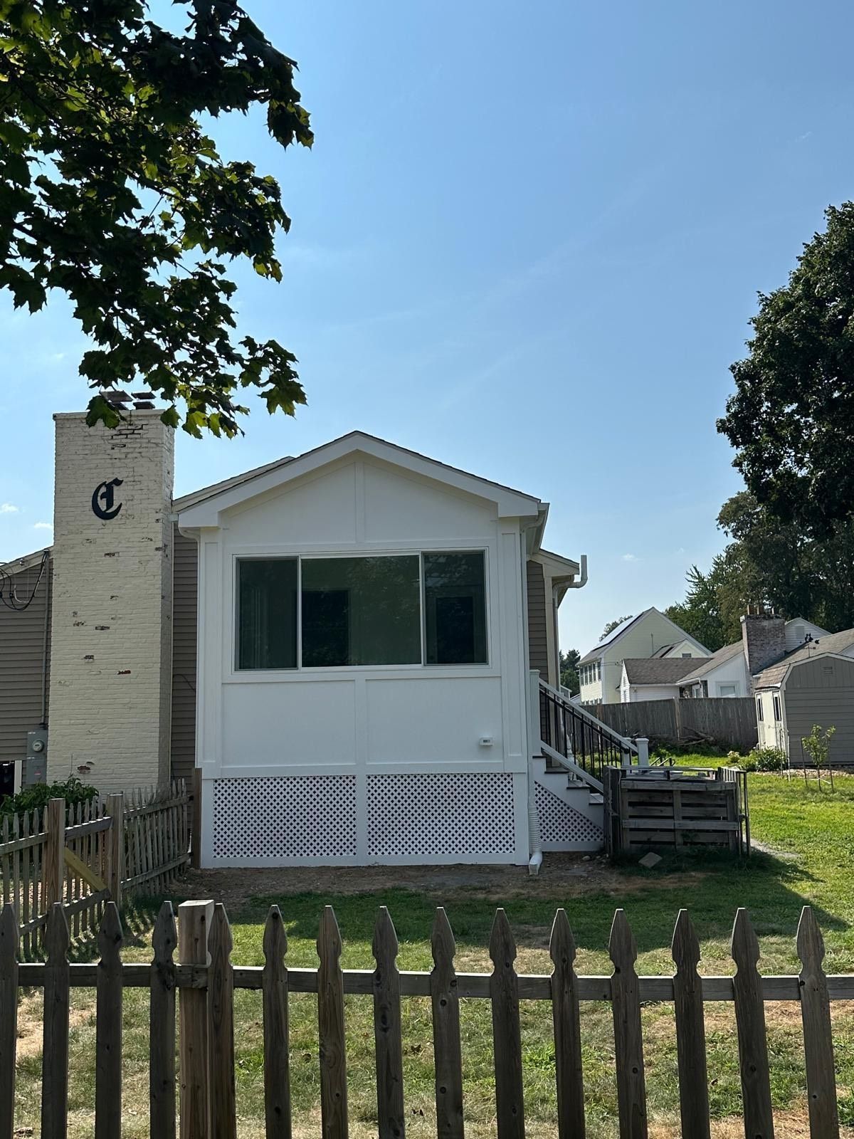 Back of a house with a white addition, brick chimney, and a wooden fence under a blue sky.