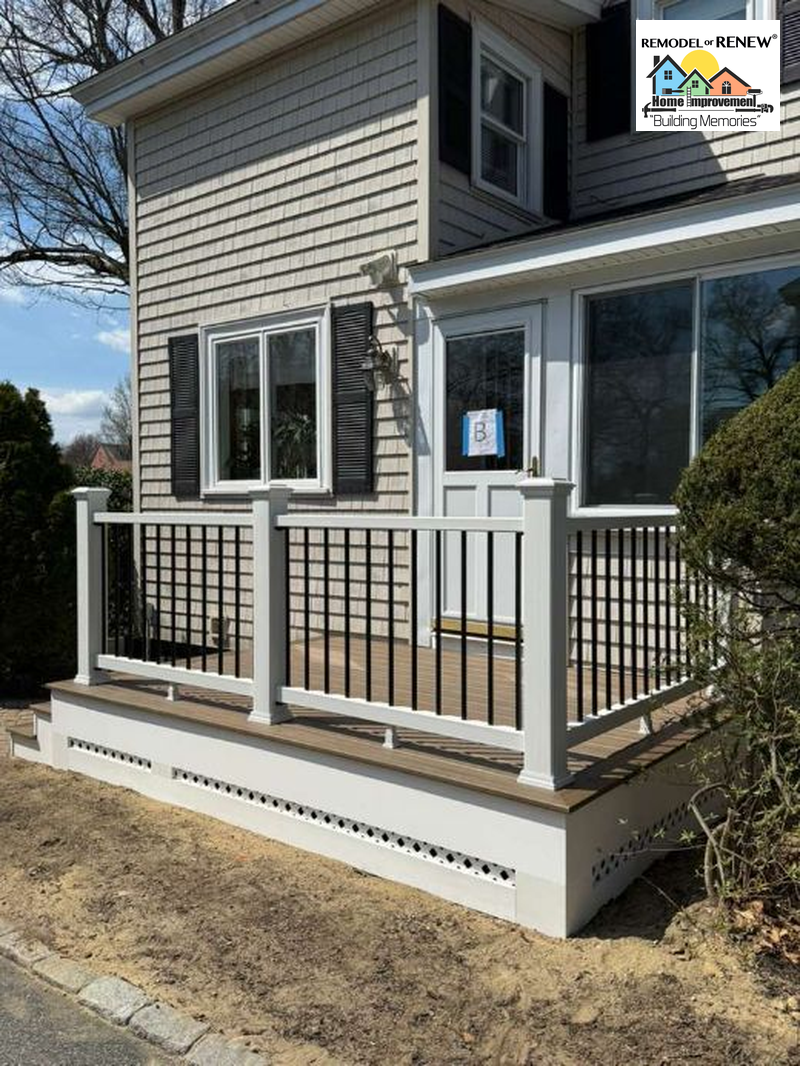 A newly constructed deck with white railings and black balusters attached to a tan house with a white door and windows.