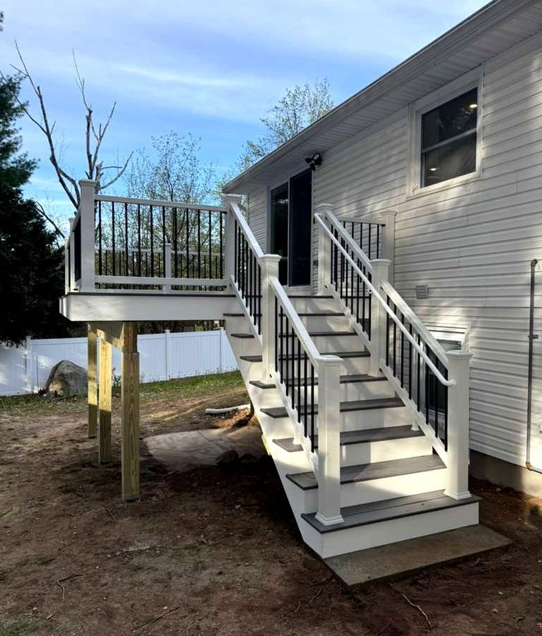 Side-view of a raised white house deck with stairs, railing, and support posts overlooking a yard and pond