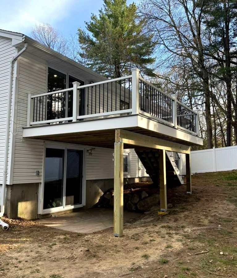 Raised deck with white railing and stairs on the side of a house, supported by posts over a sloped yard