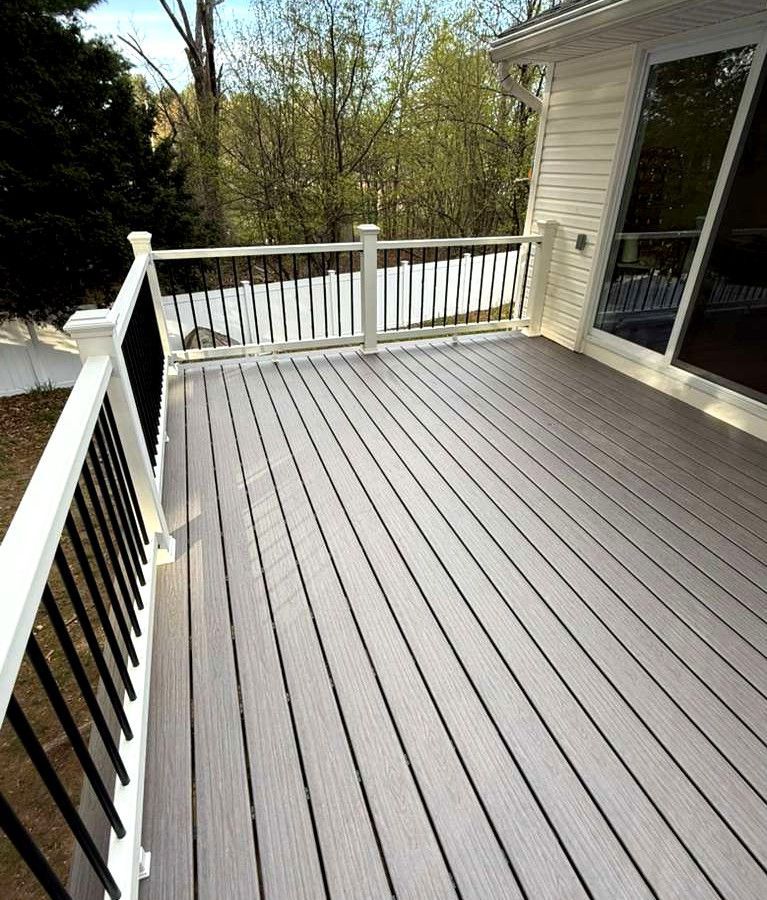 White wooden deck with railing beside a house, overlooking trees and snow outside.